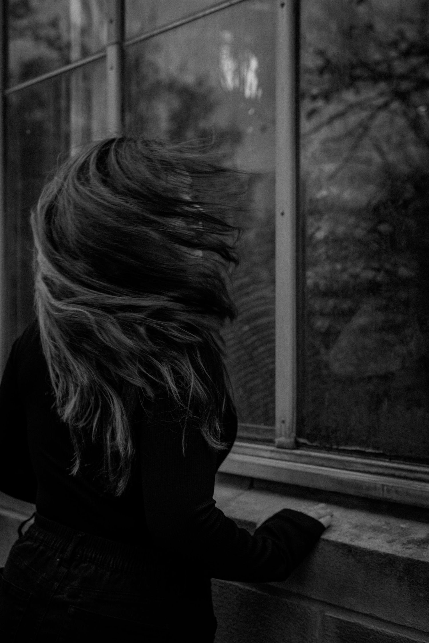 Black and white photo of woman tossing hair near window with trees reflected in glass.