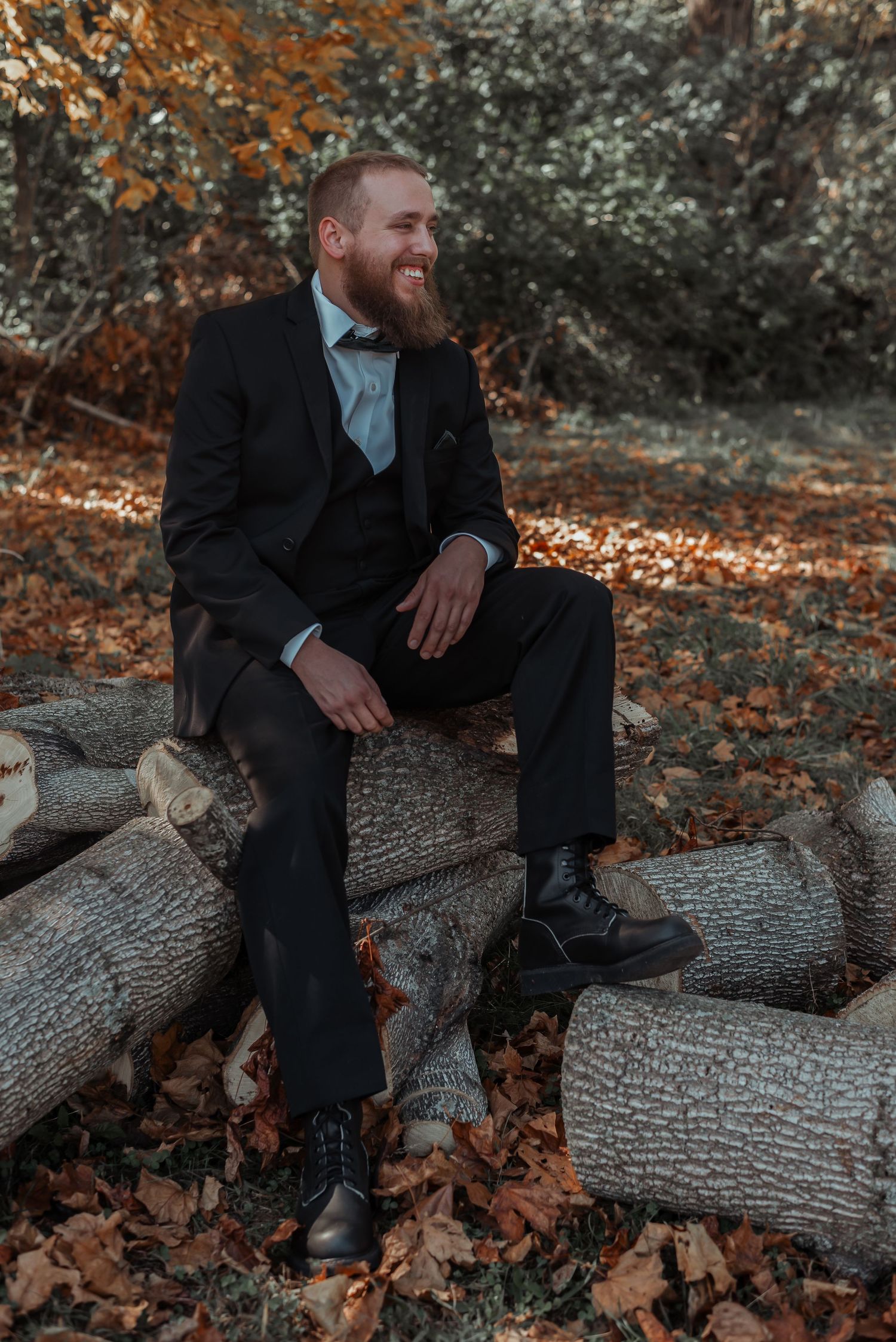 Bearded groom in a black suit smiles while sitting on logs surrounded by autumn foliage outdoors.