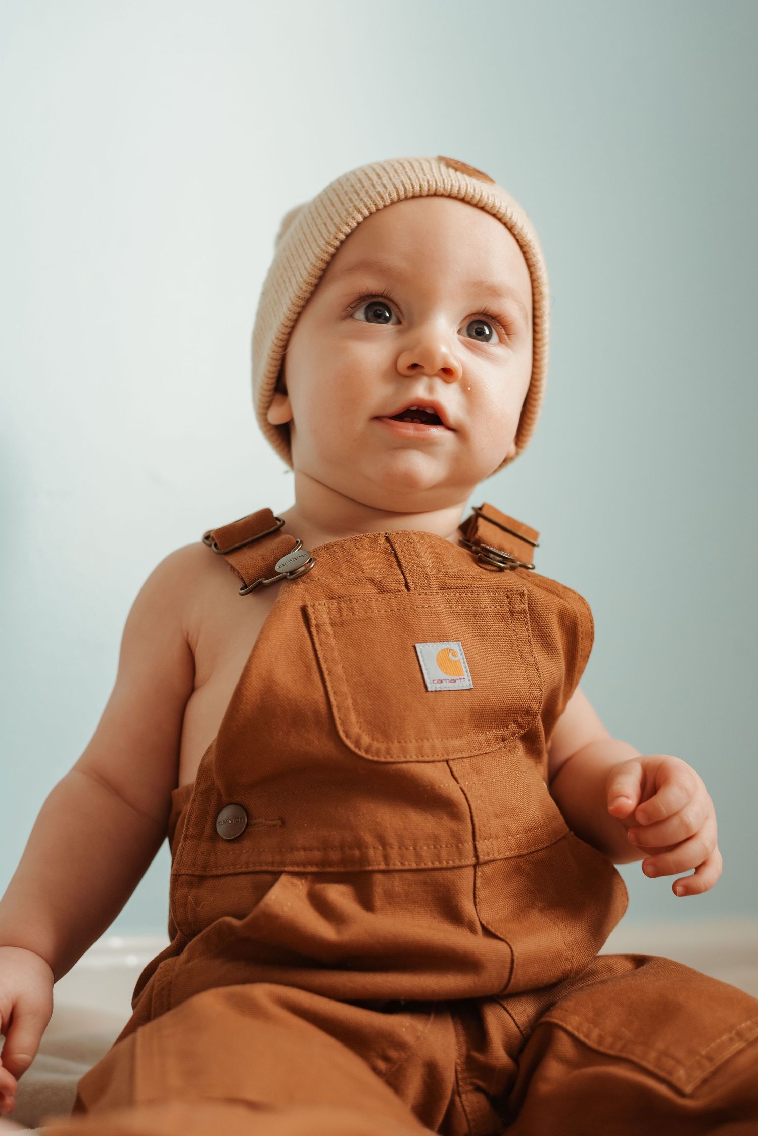 Cute baby wearing brown Carhartt overalls and cream knit beanie sits on light background.