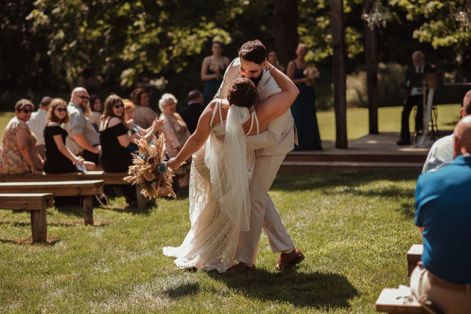 A couple shares a romantic dip kiss during an outdoor wedding ceremony on a sunny green lawn.
