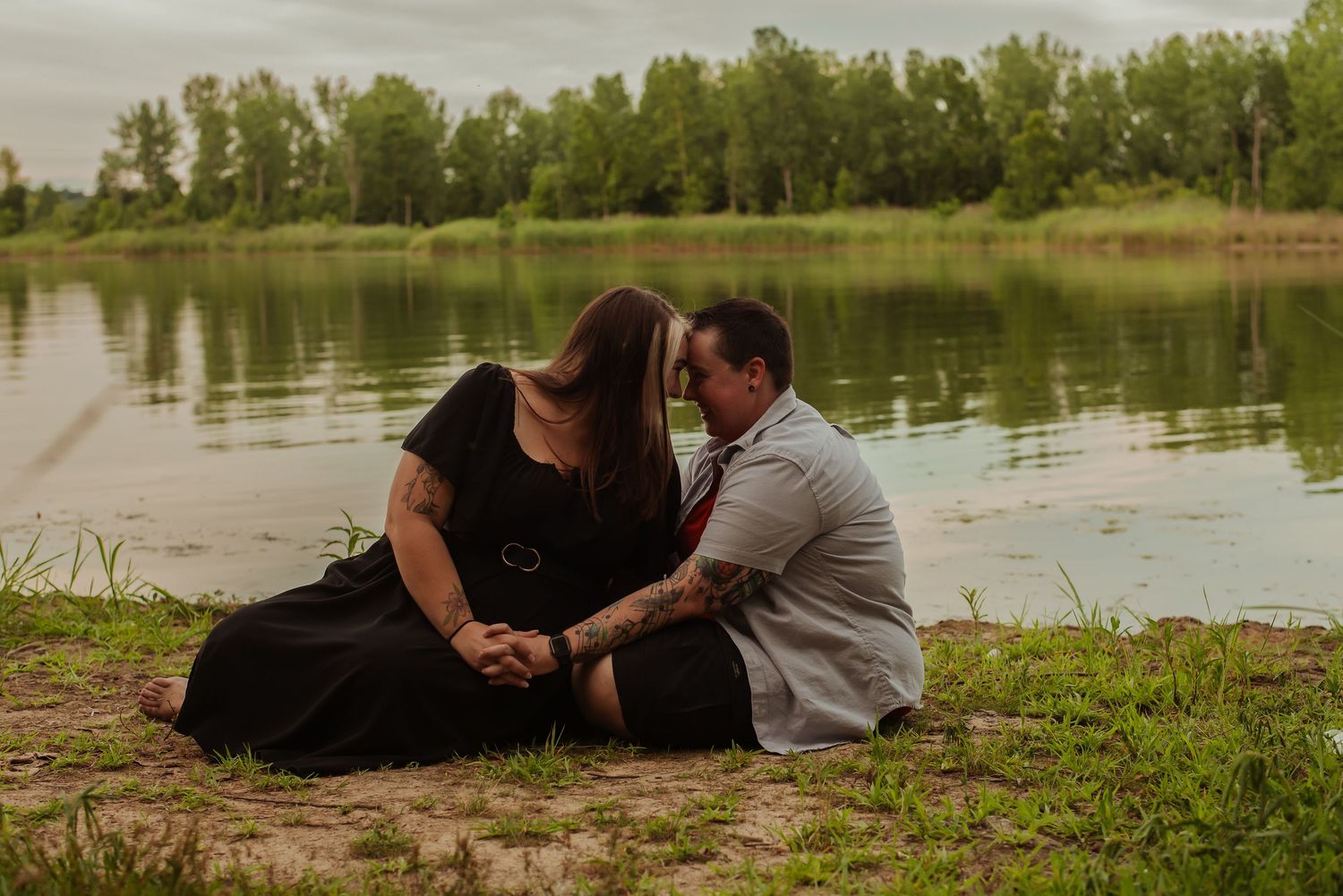 Couple sitting close together near a serene lake shore, surrounded by lush greenery, sharing a romantic moment outdoors.