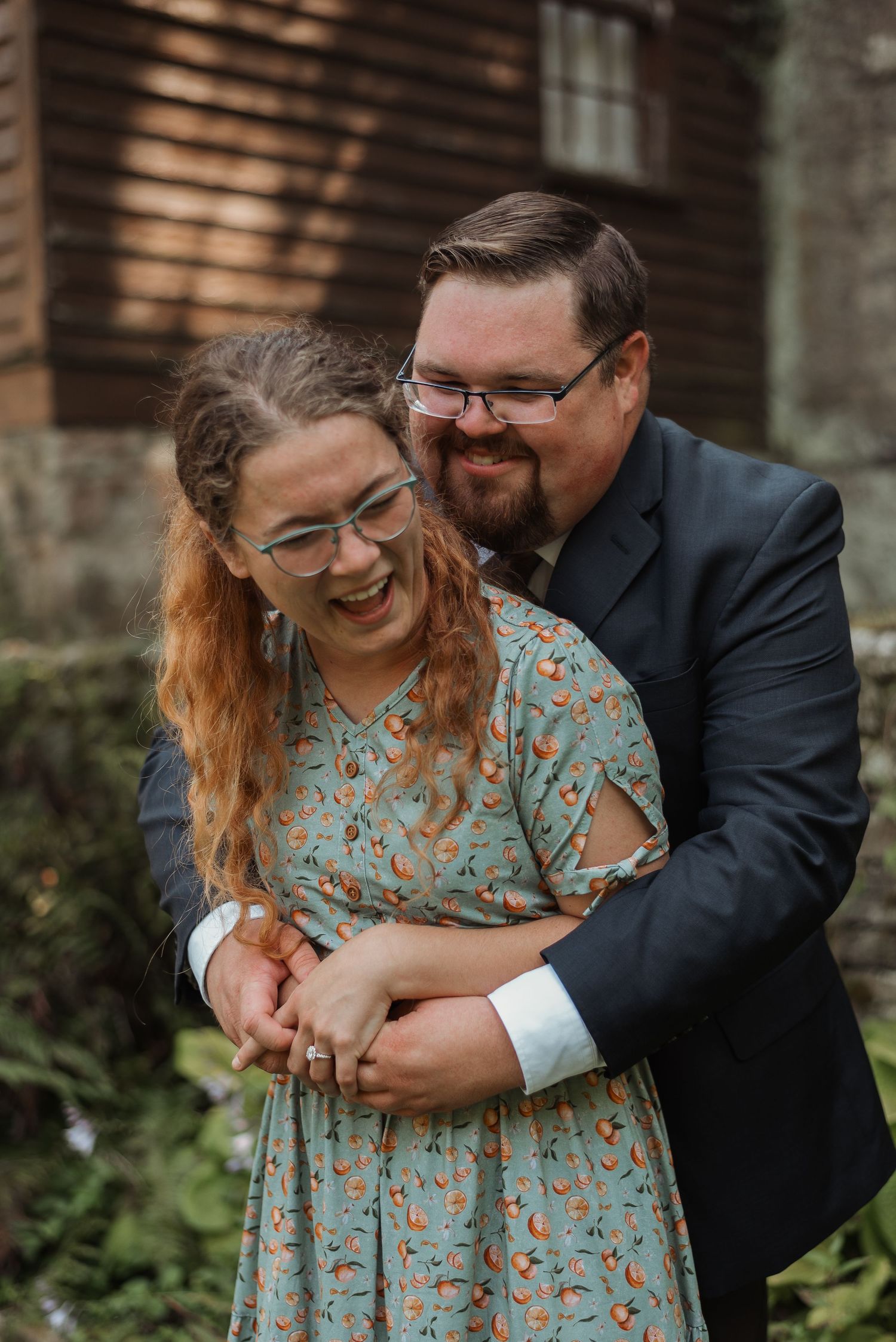 A laughing woman in a floral dress is embraced from behind by a smiling bearded man in a suit.
