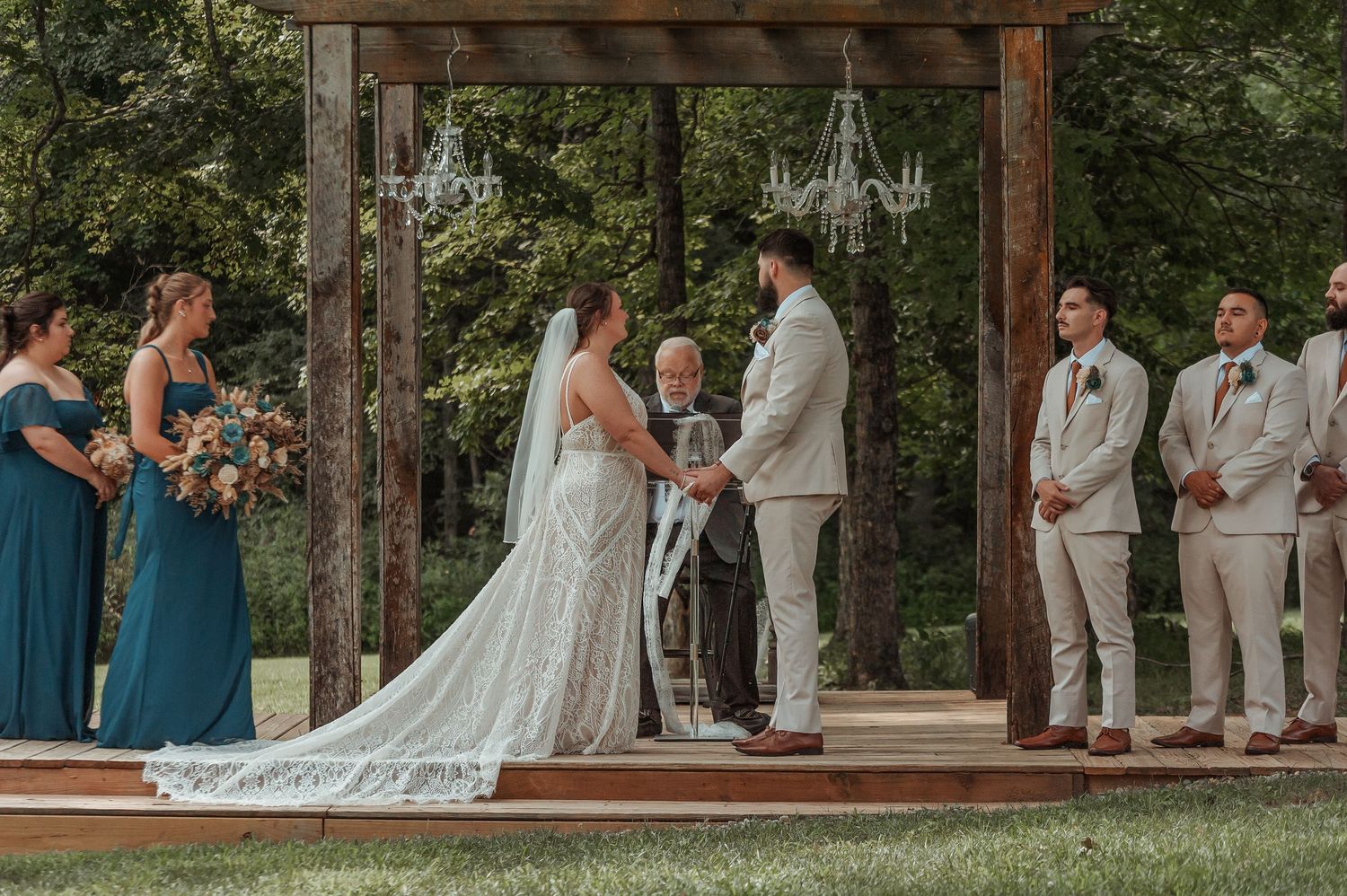 Bride and groom exchange vows under a wooden pergola with chandeliers surrounded by wedding party outdoors.