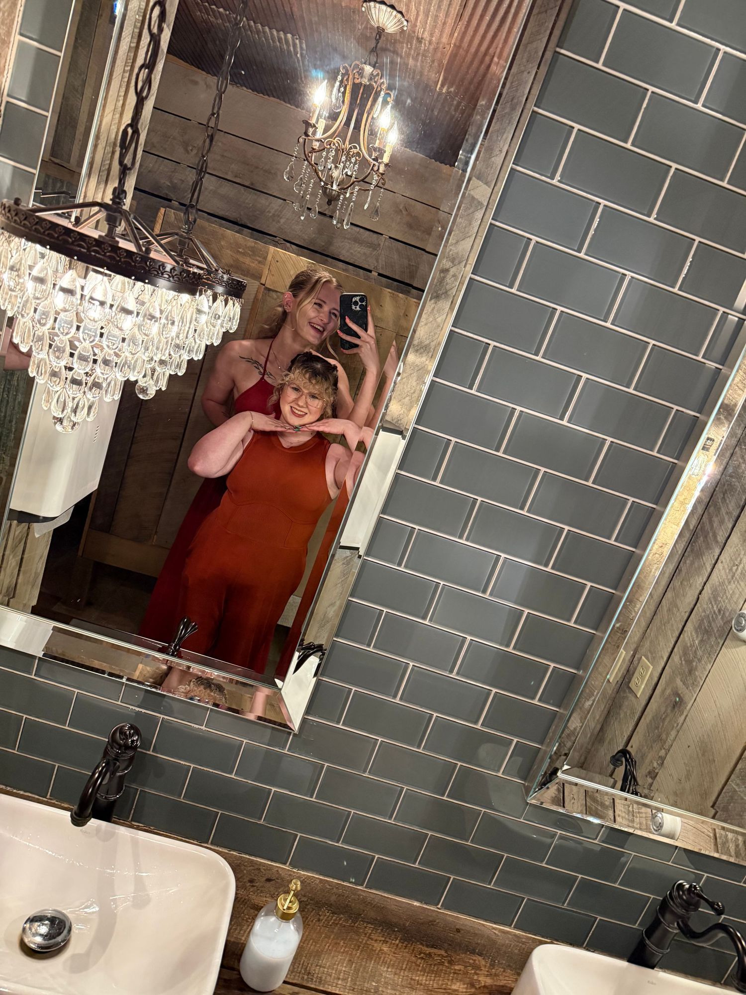 Two women in elegant dresses taking a mirror selfie in a stylish tiled bathroom.