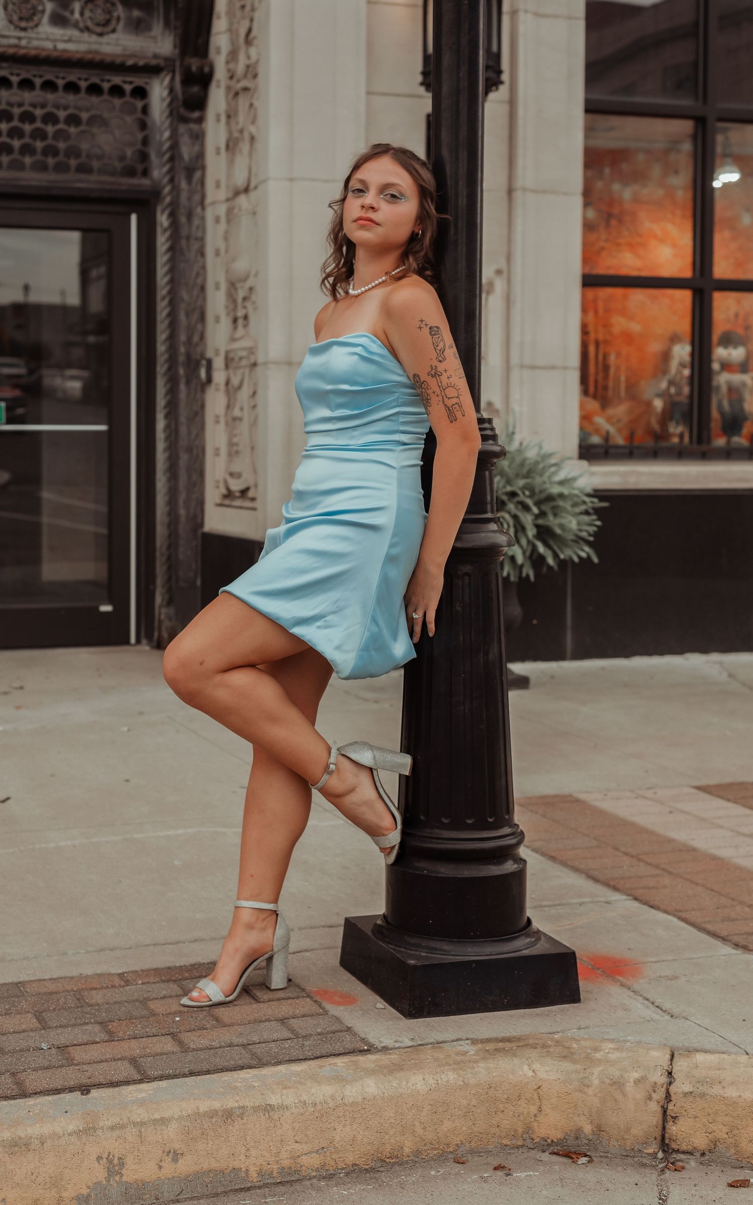 Woman in light blue strapless mini dress leaning against black street pole on urban sidewalk.
