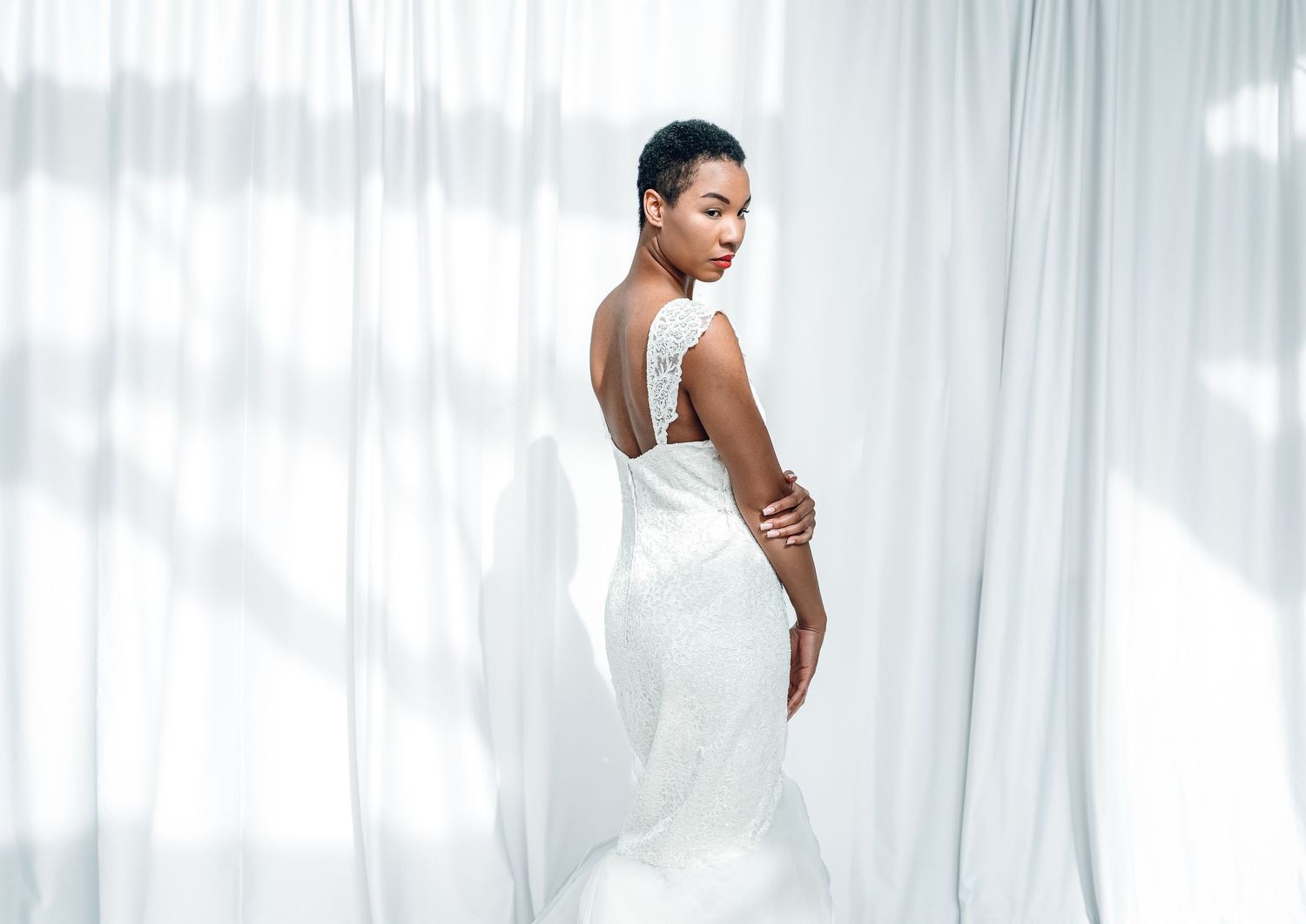 Elegant bride in fitted white lace gown posing gracefully against flowing white curtains.