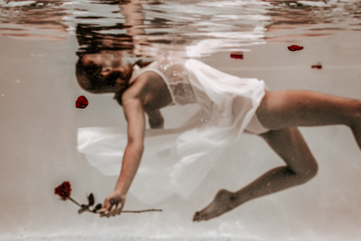 Woman in white dress reaching for red rose underwater with floating petals around her.