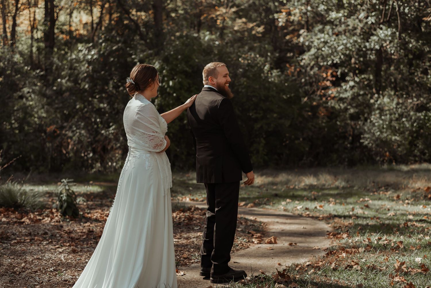 Bride in white dress approaching groom from behind in wooded setting.