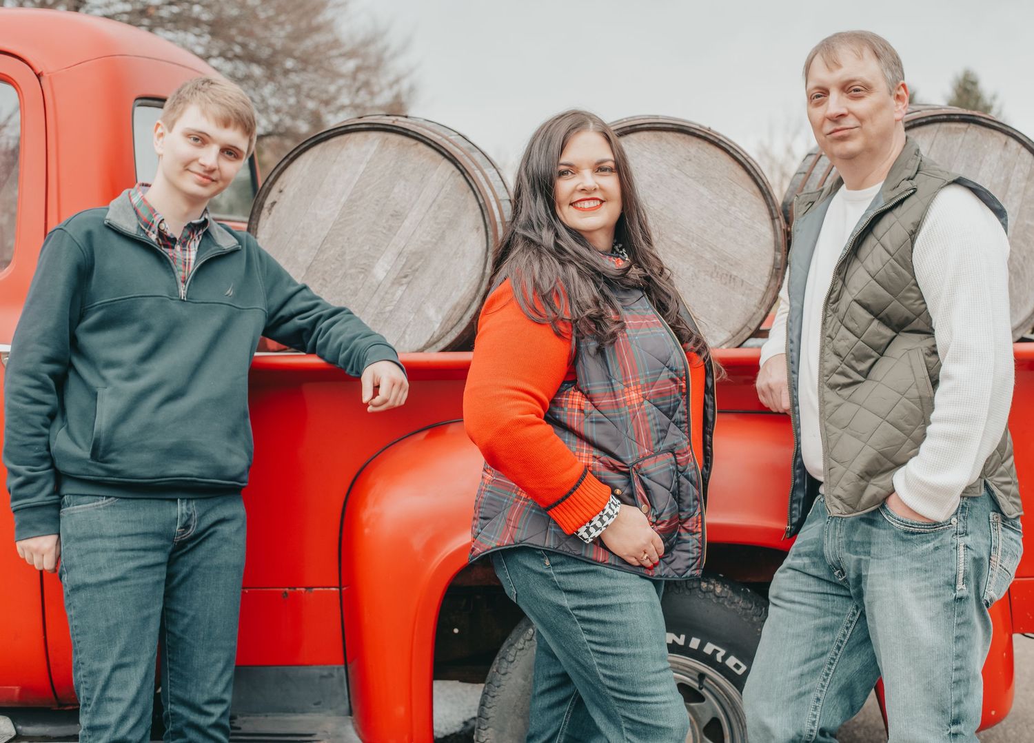 Family of three smiles leaning against a vintage red truck with wooden barrels during outdoor portraits.