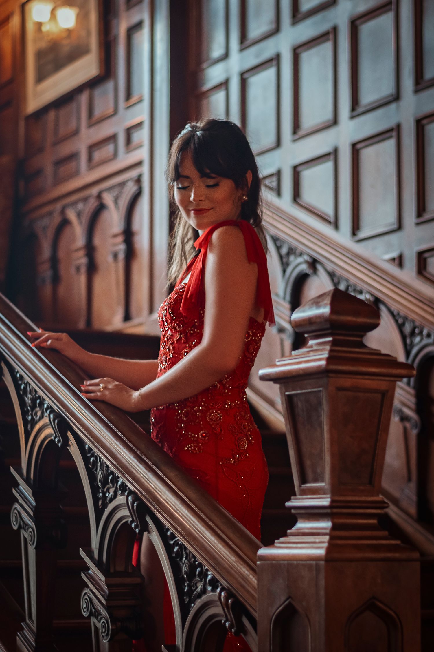 Woman in red embellished gown looking down while holding a carved wooden staircase railing.