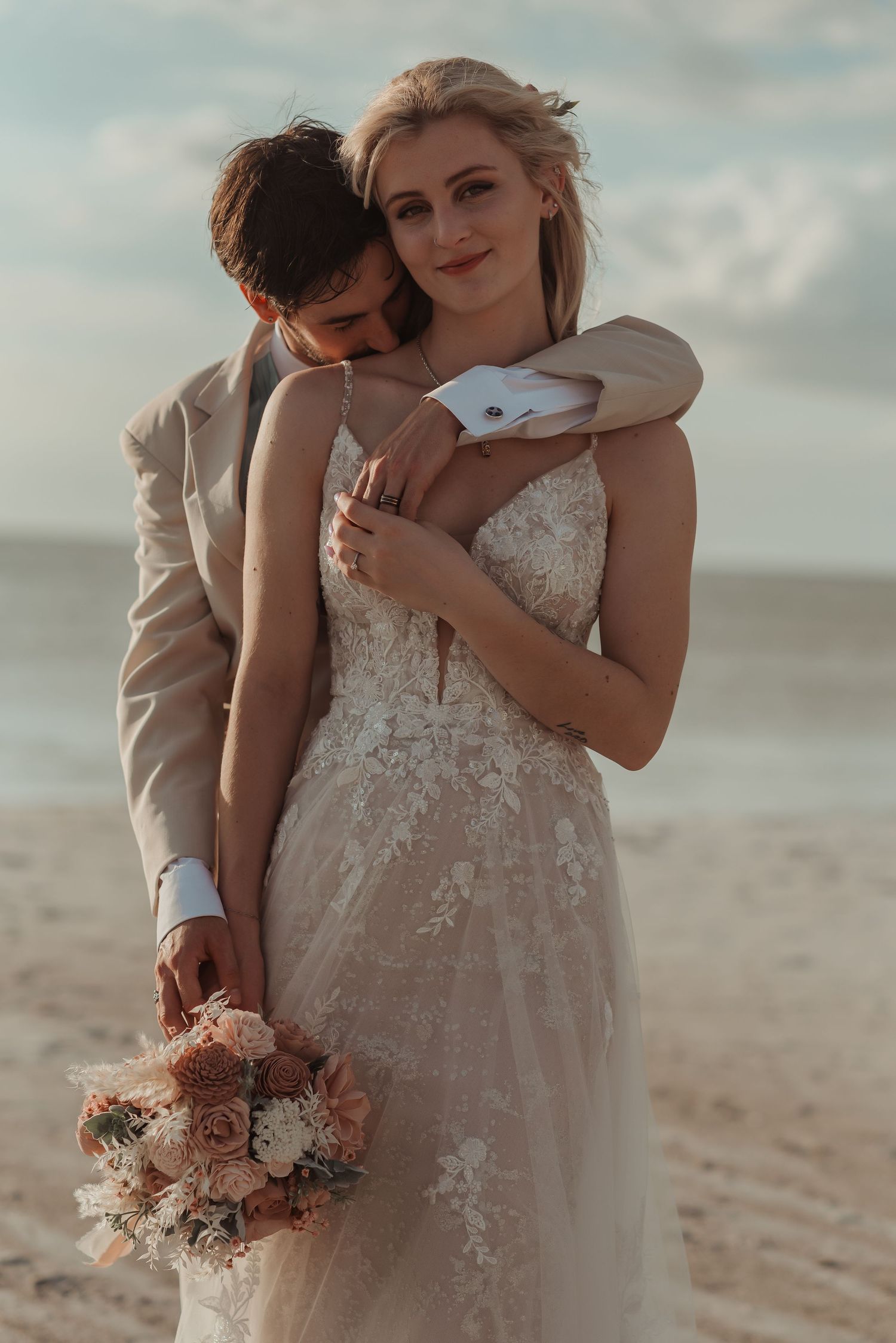 Groom embracing bride from behind on beach as she holds blush pink and white bouquet.