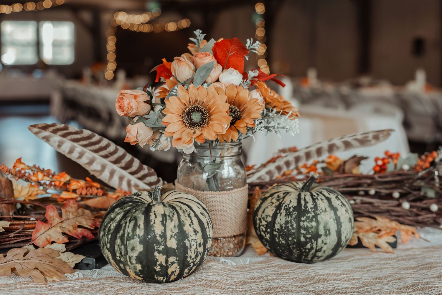 Fall wedding centerpiece with orange sunflowers, pumpkins, feathers, and autumn leaves on a table.