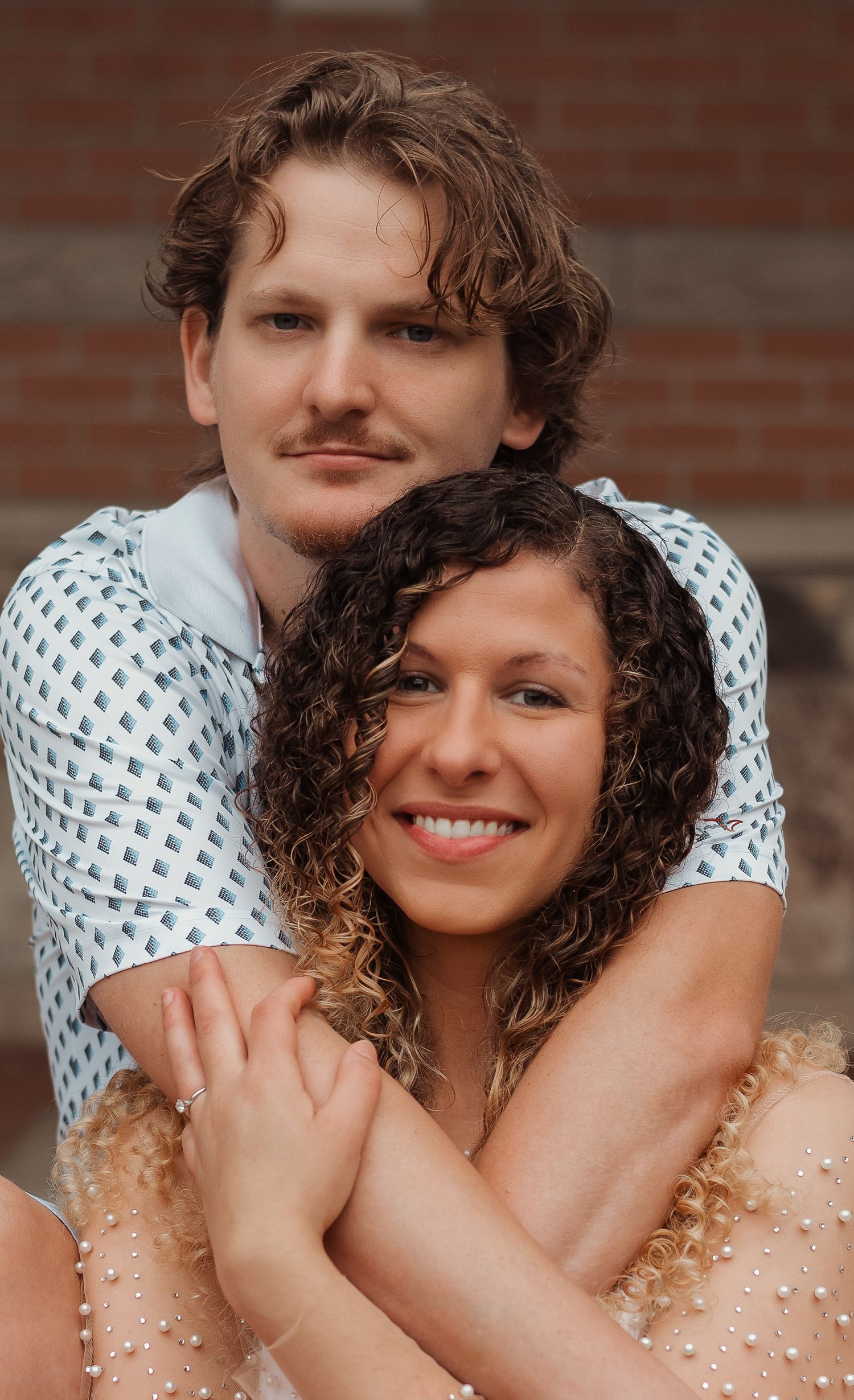 Smiling couple with curly hair posing together in front of a brick wall backdrop.