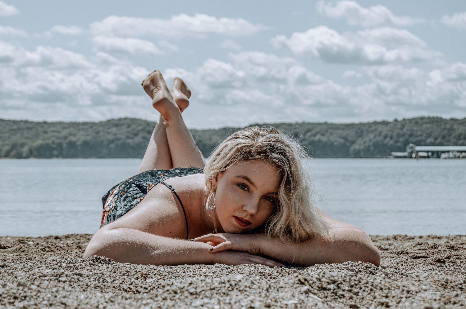 Blonde woman in floral swimsuit lying on a sandy beach by the lake.