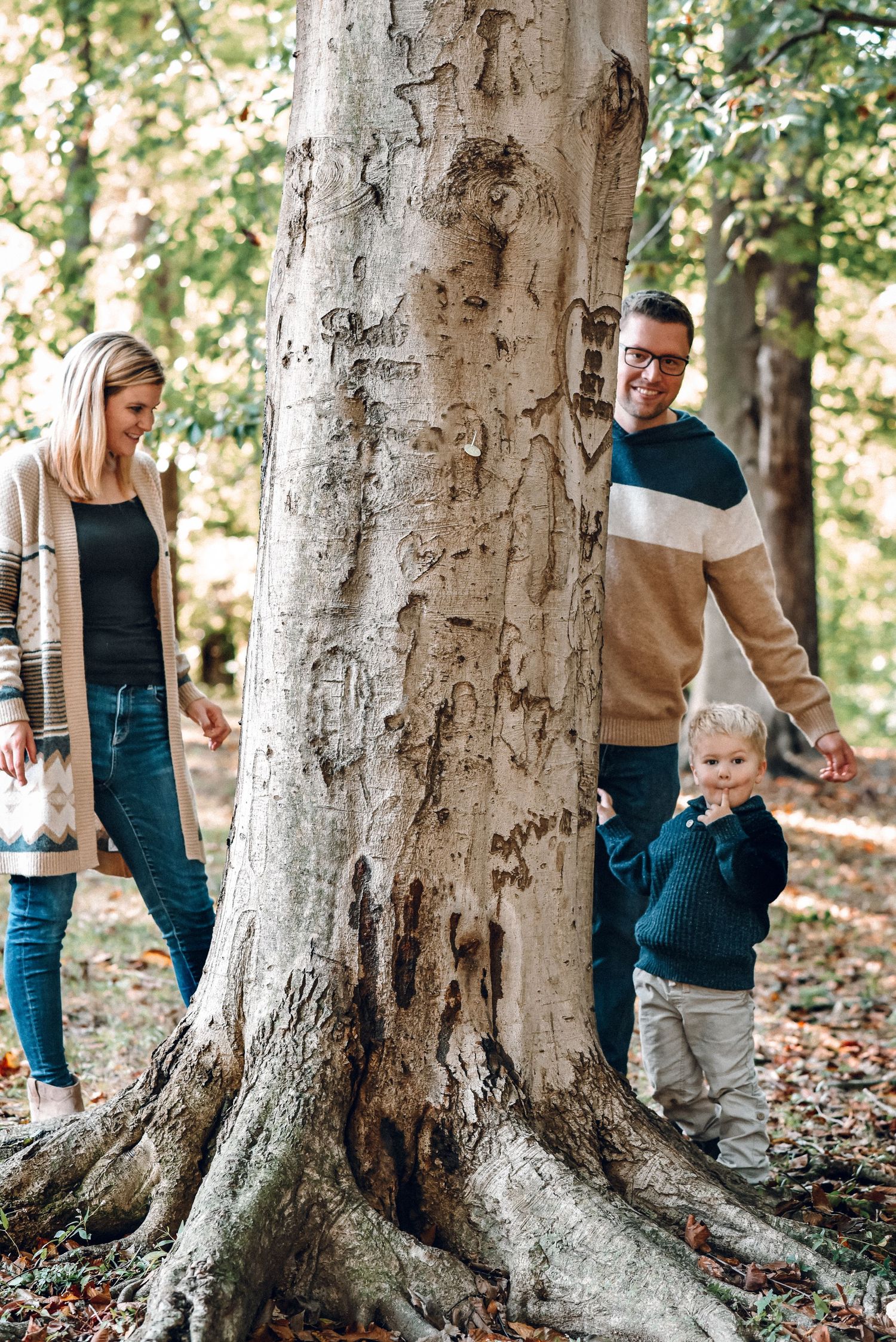 Young family with toddler explores around a large tree trunk during an outdoor fall family photo session.