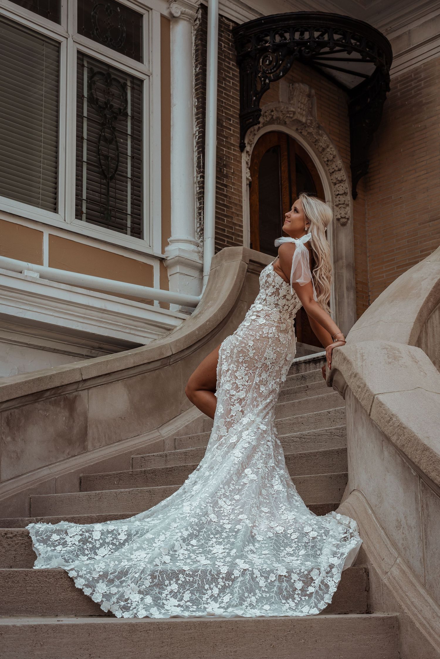 Bride in floral lace wedding gown with train posing on ornate stone staircase of historic building.