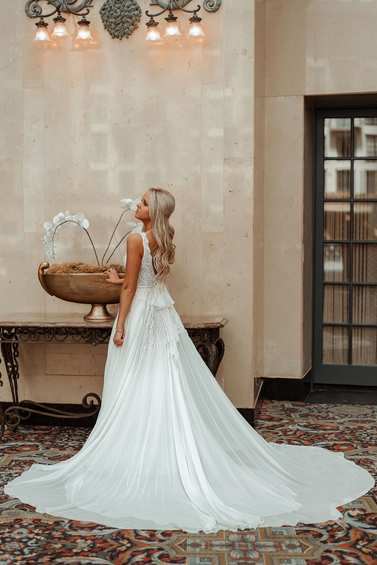Bride in sleeveless lace gown with dramatic flowing train poses in elegant venue with ornate decor.
