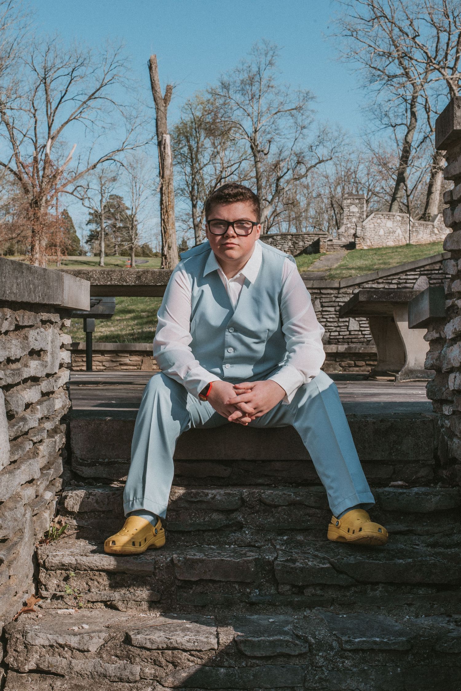Young man in light blue suit and yellow Crocs sits on stone steps outdoors at prom.