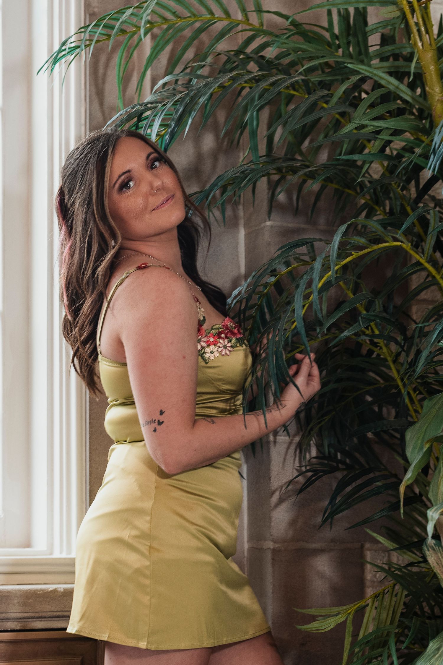 Young woman in yellow floral cami dress posing beside a tropical palm plant by a window.