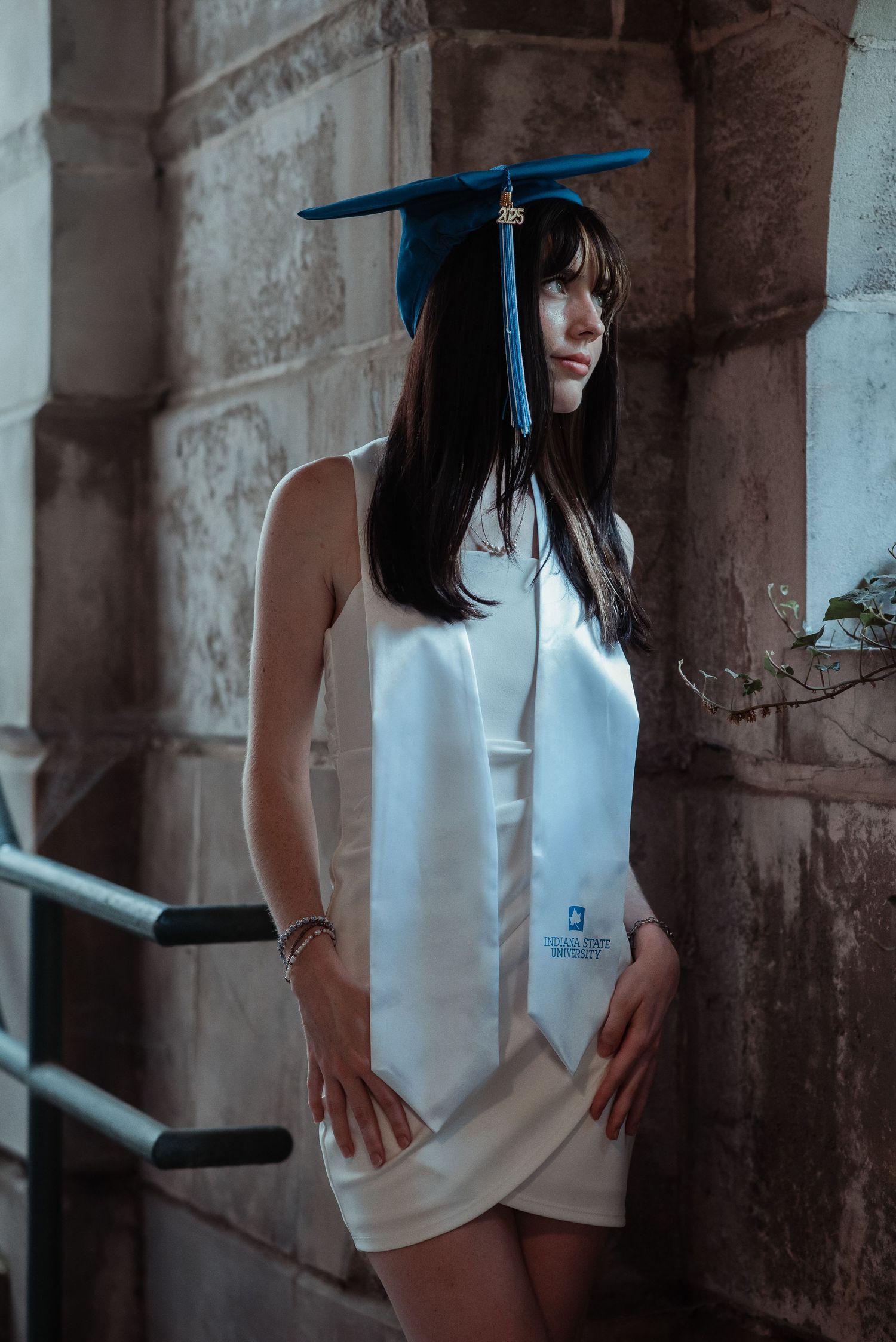 Graduate in blue mortarboard cap and white honor stole poses against stone wall in elegant white dress.