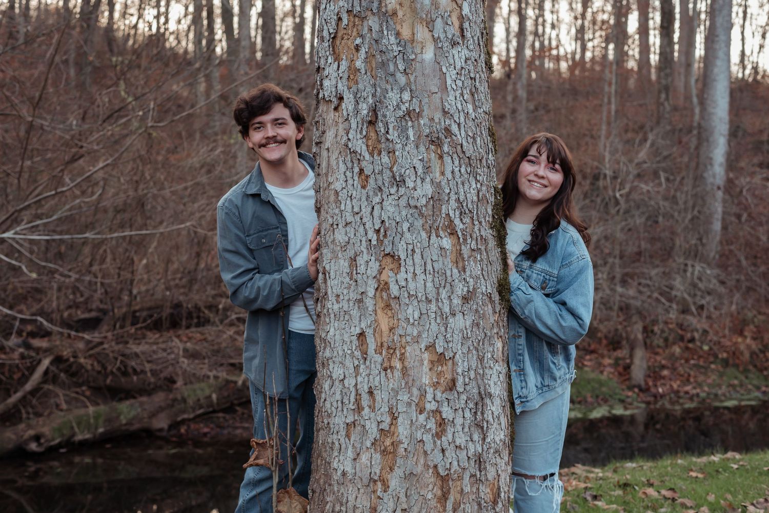 A smiling couple peeks out from behind a large tree trunk in an autumn forest setting.