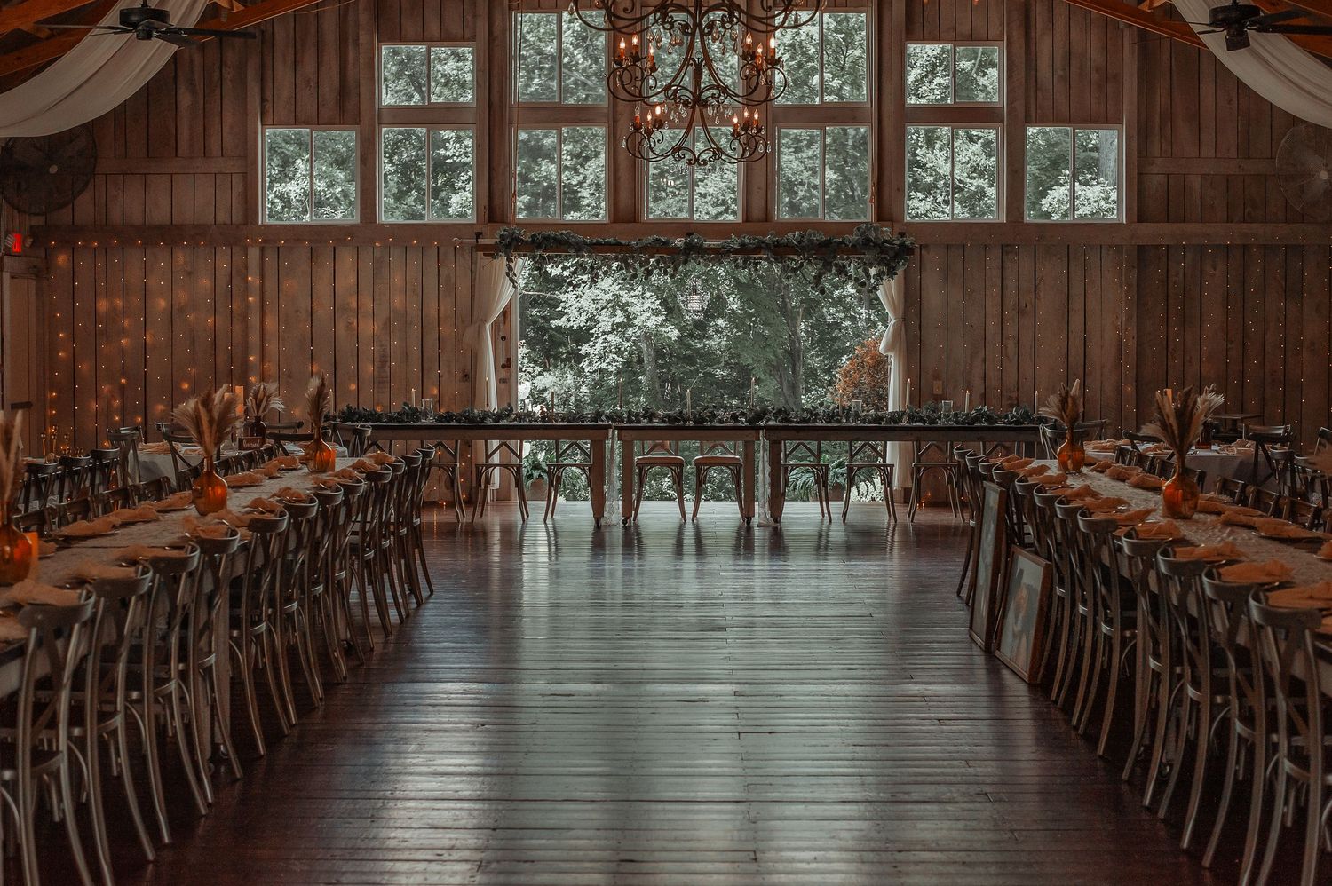 Elegant rustic barn wedding reception hall with long tables, cross-back chairs, and chandelier lighting.