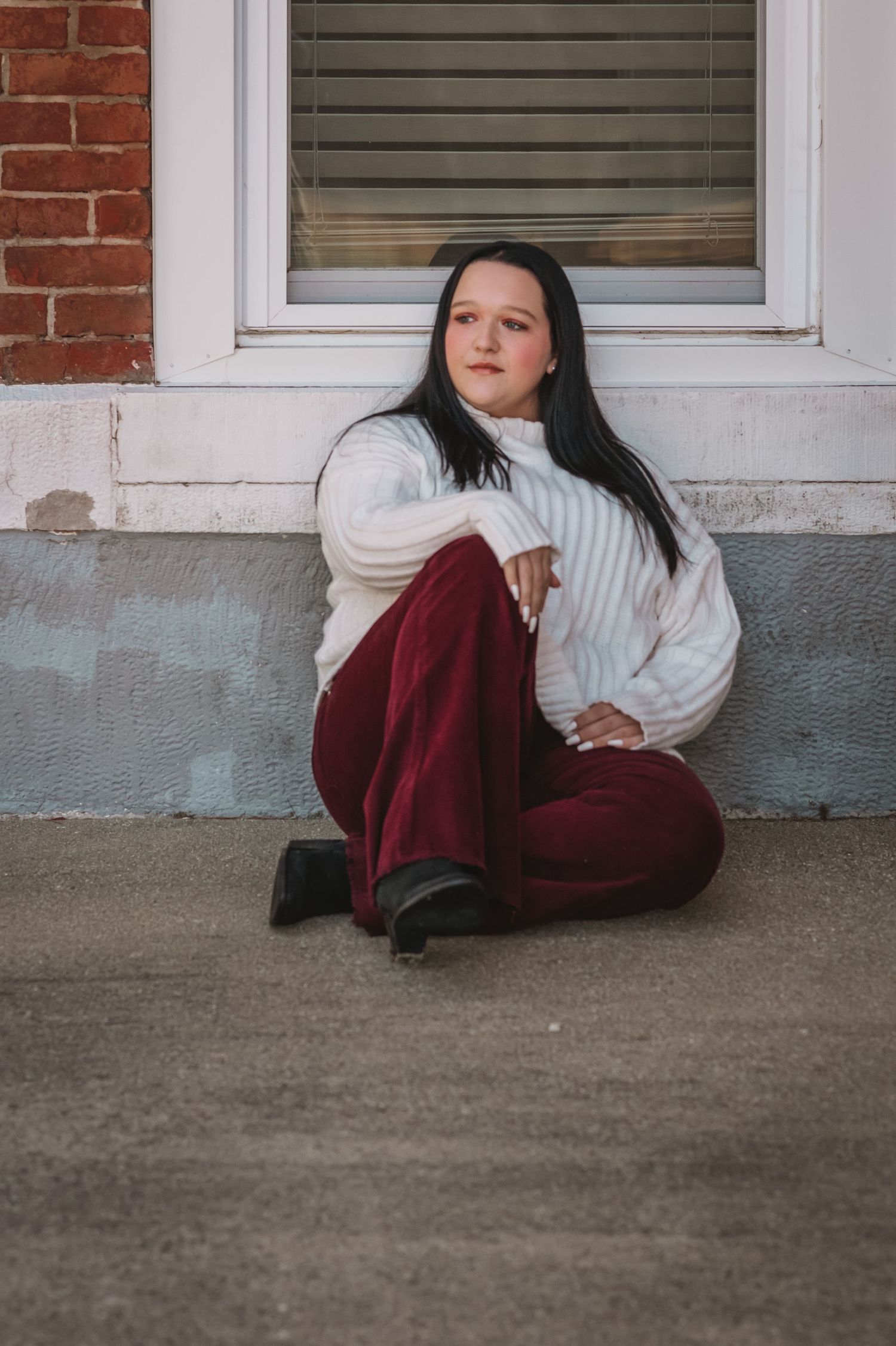 Young woman with dark hair in white sweater and burgundy pants sits cross-legged on sidewalk.