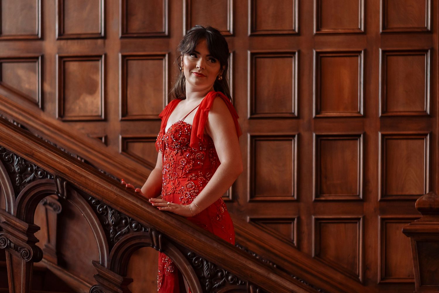 Woman in red sequin halter gown posing on carved wooden staircase against paneled wood wall.