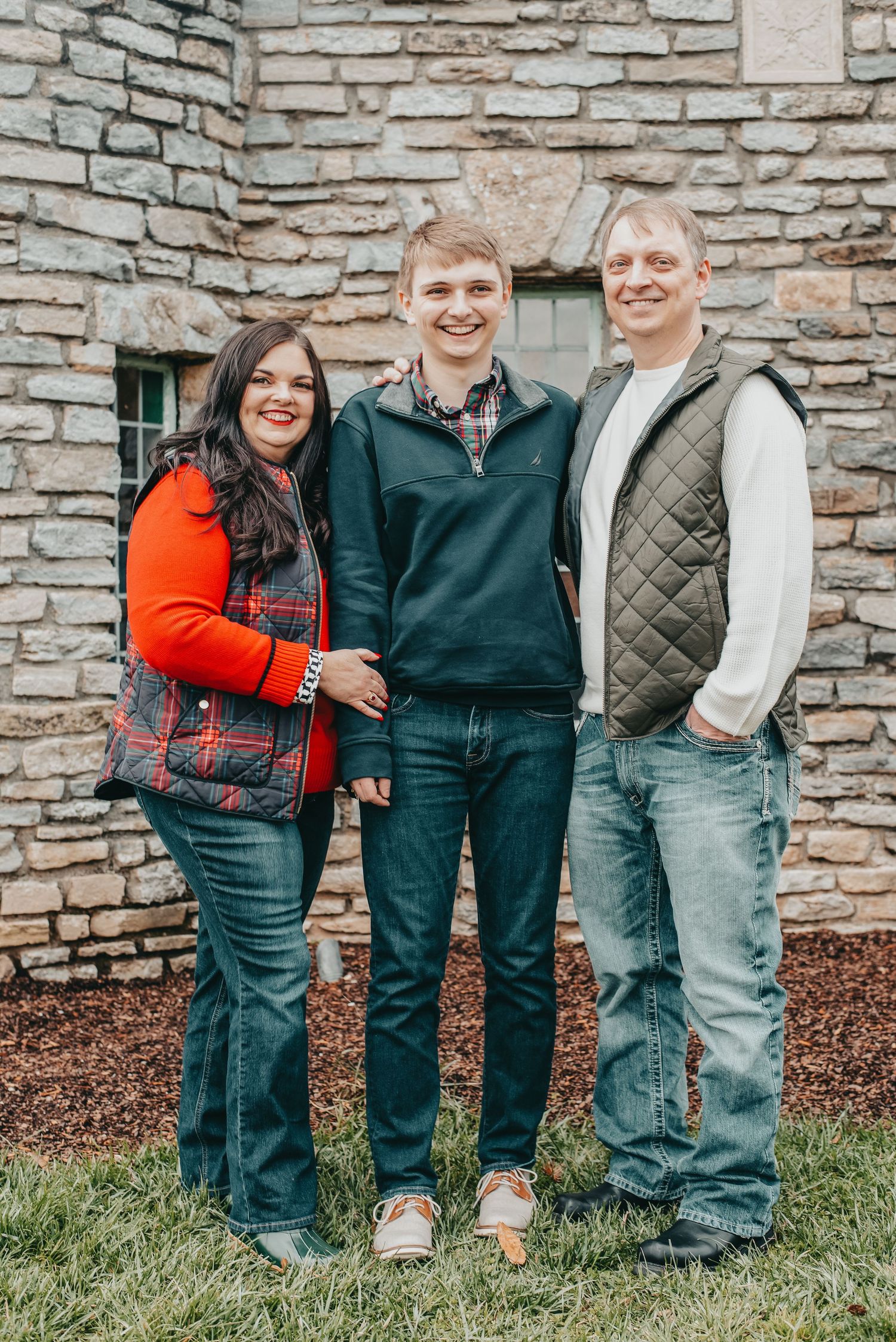 Family of three smiling together outdoors in front of a stone building, wearing casual fall outfits.