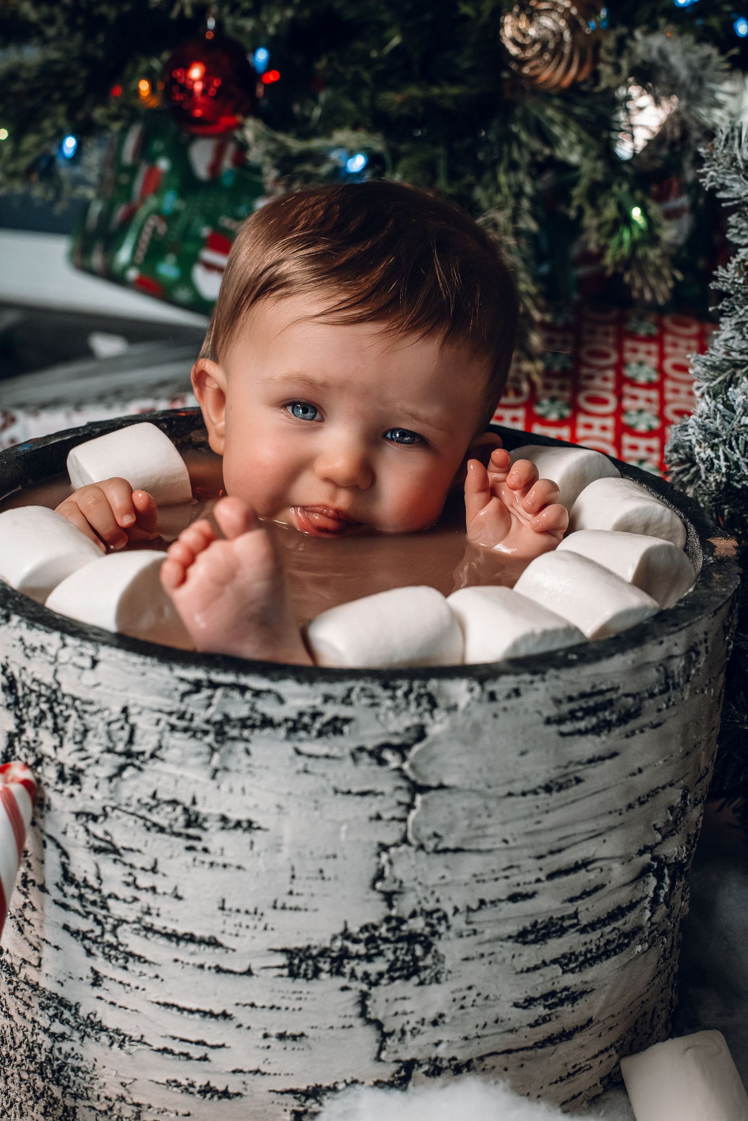 Happy baby with blue eyes sits in marshmallow-filled barrel during festive Christmas photo session.