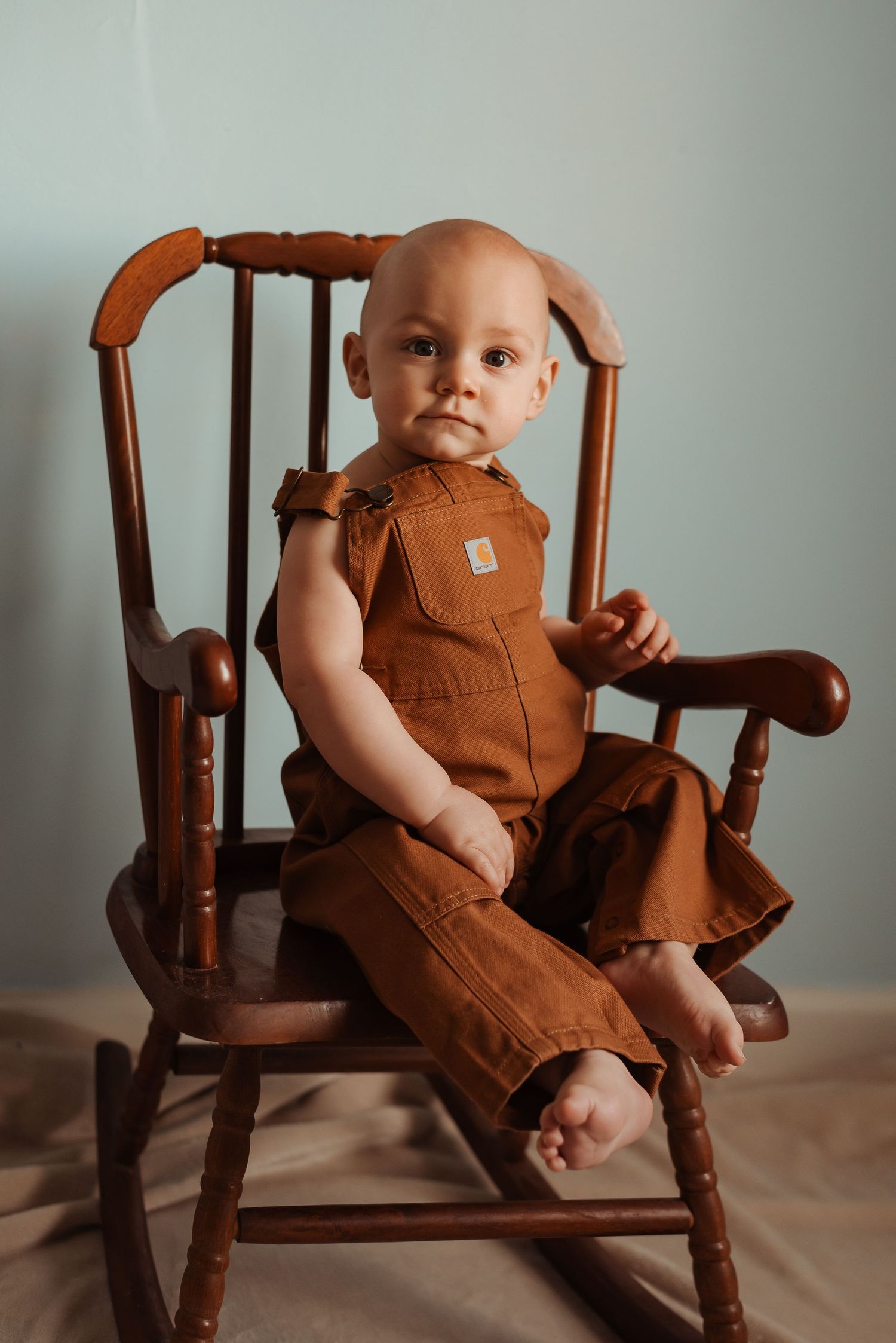 Adorable baby in brown Carhartt overalls sits in a wooden rocking chair on a neutral background.
