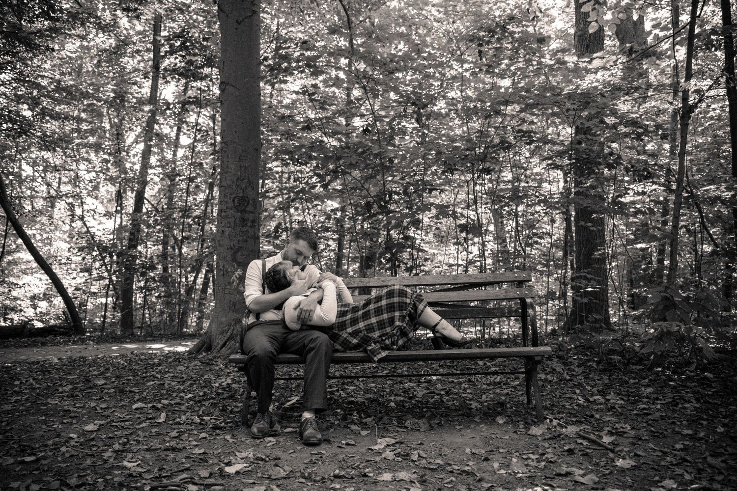 Black and white photo of a couple sitting closely together on a park bench surrounded by fallen autumn leaves.