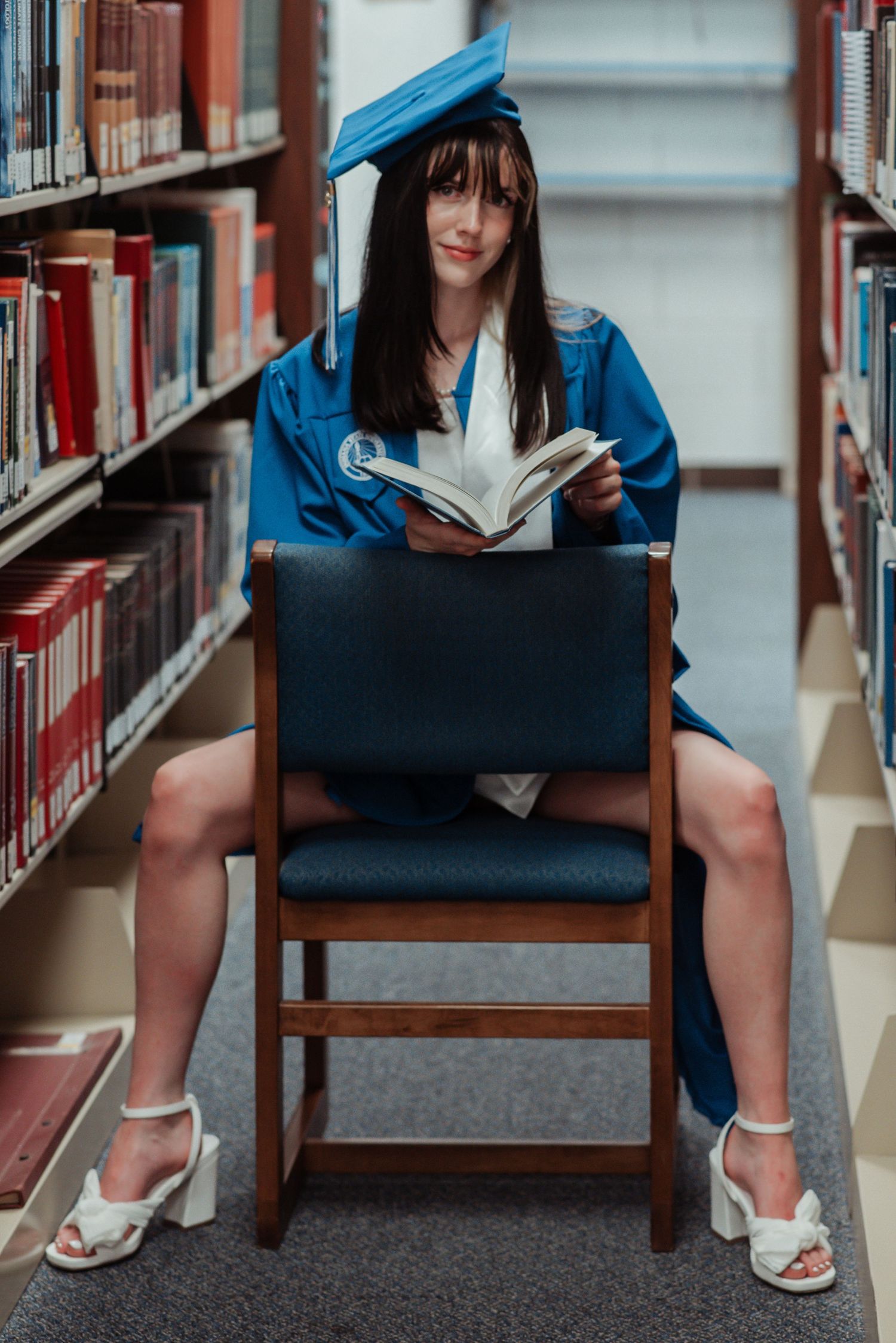 Graduate in blue cap and gown sits in library chair reading book surrounded by bookshelves.