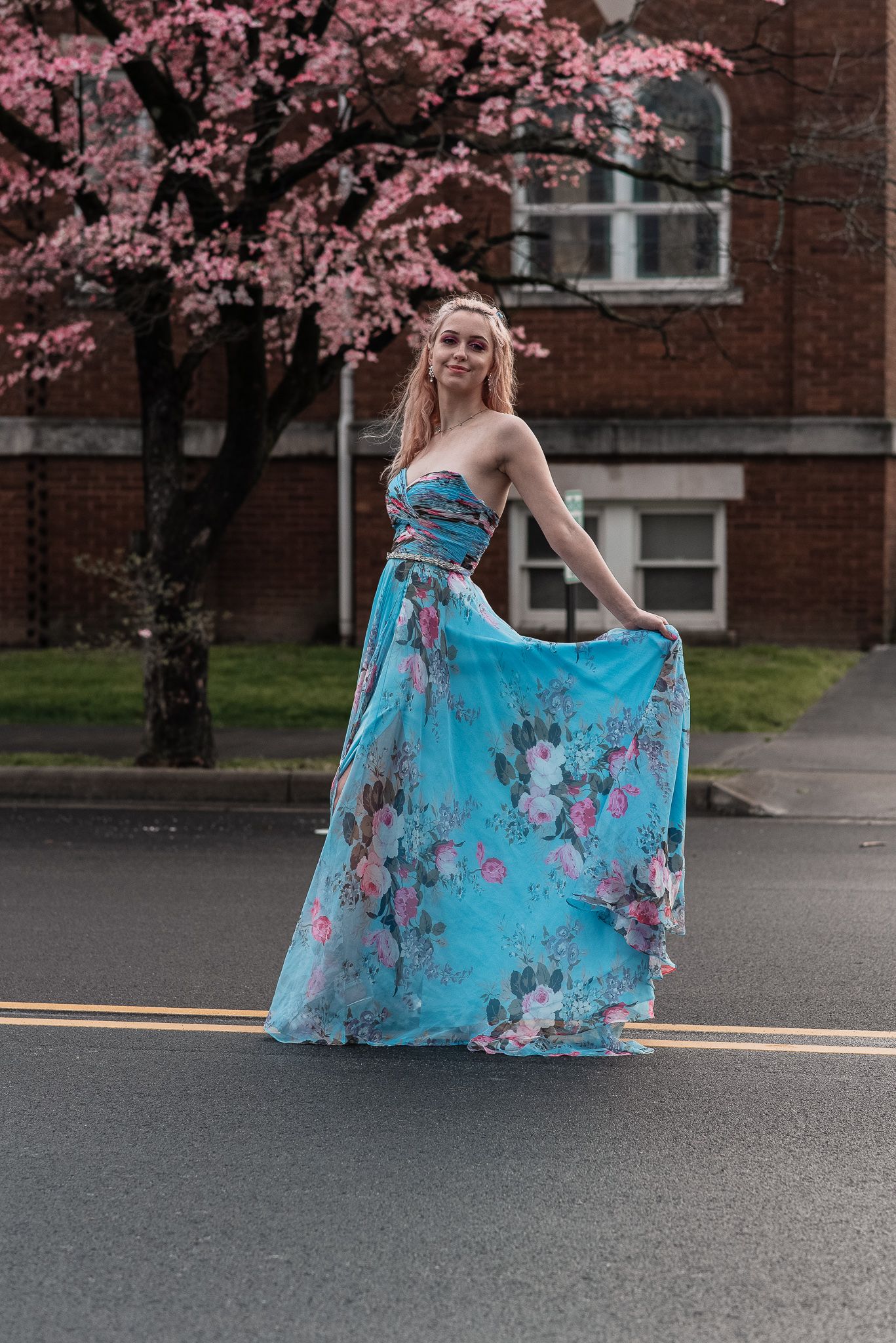 Young woman in flowing blue floral strapless gown twirling on street near pink cherry blossom tree.
