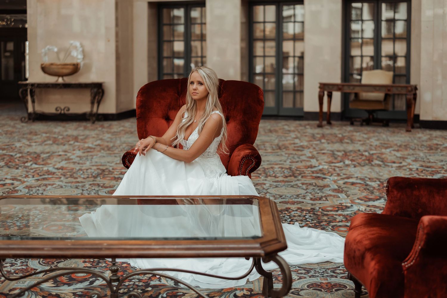 Elegant bride in white wedding gown leaning on a glass coffee table surrounded by red velvet furniture.