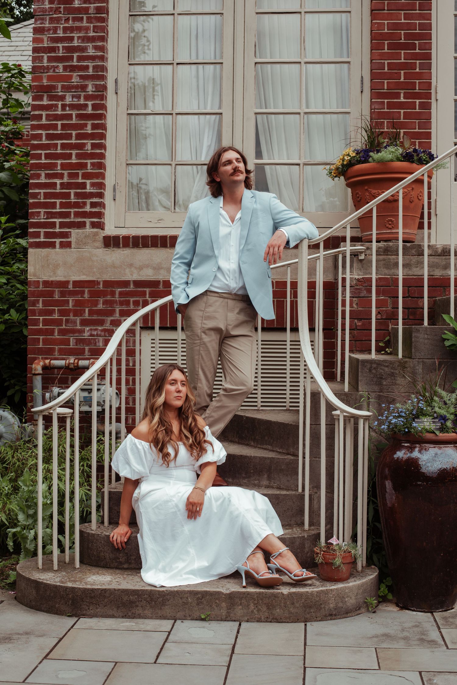 Couple posing on brick building steps, woman in white dress seated, man in light blue blazer standing behind her.
