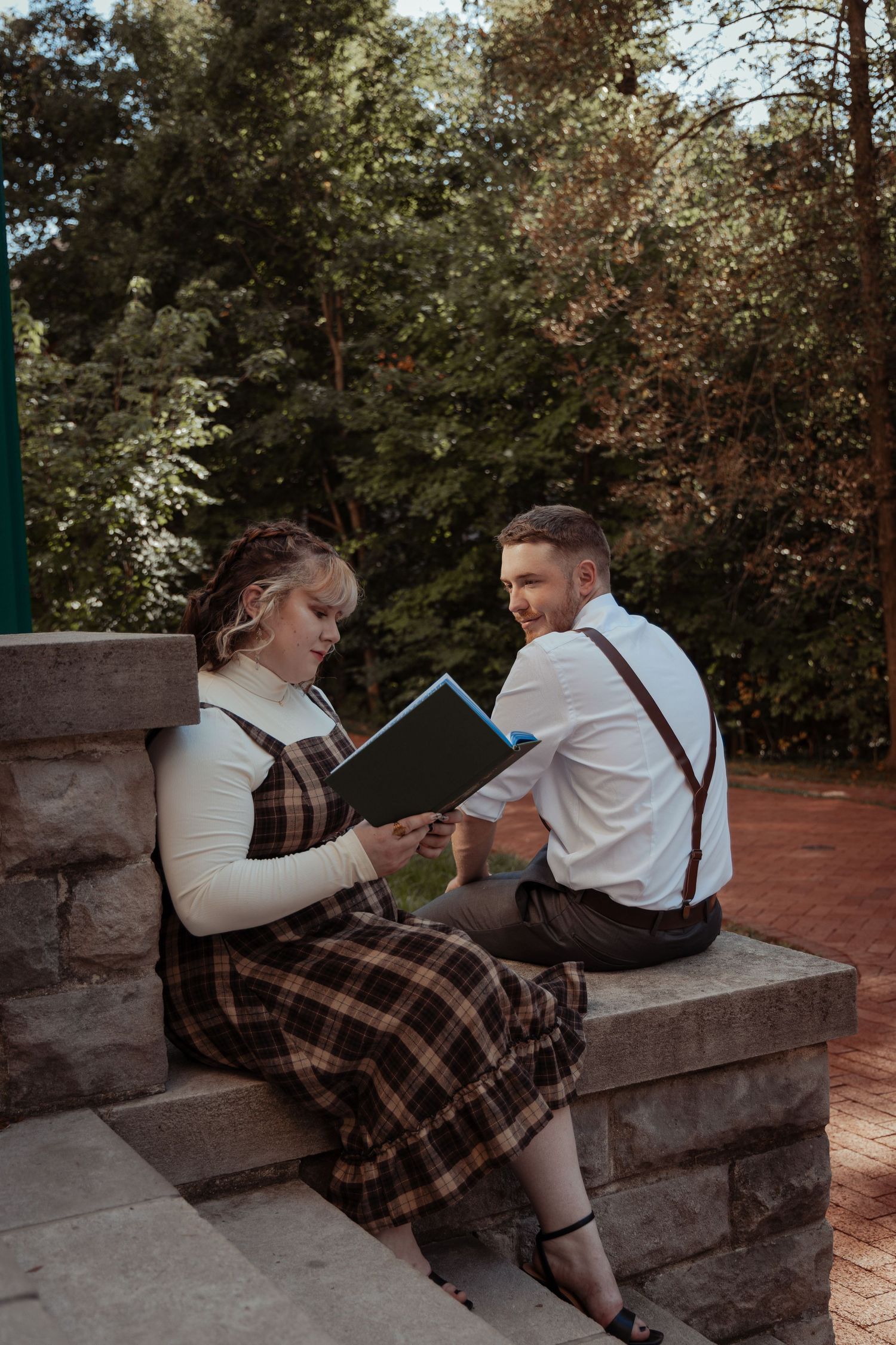 Couple sitting on stone steps in a park, woman reading a book in plaid dress, man in white shirt and suspenders.