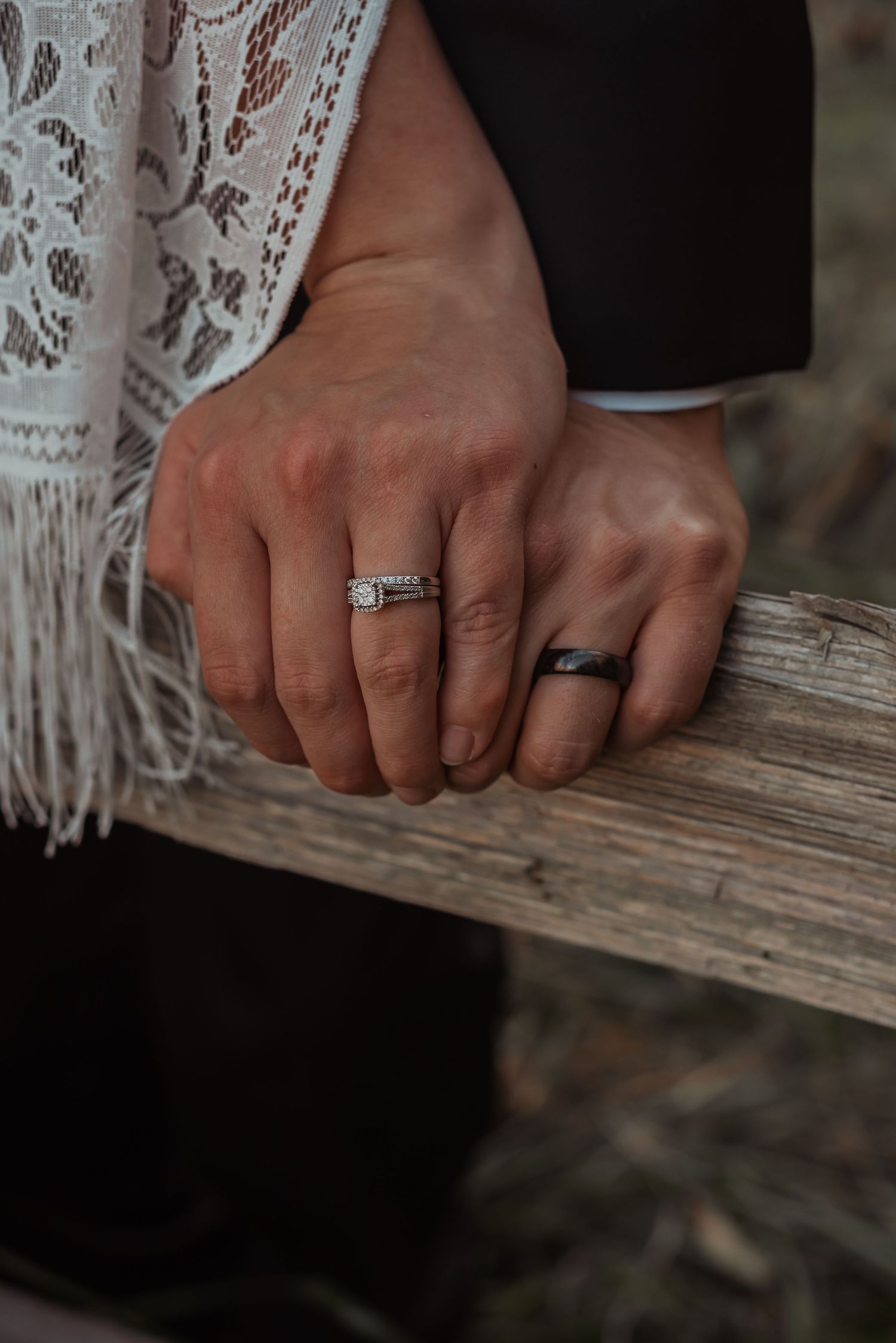 Close-up of couple's hands resting on a wooden fence, wearing matching wedding and engagement rings.