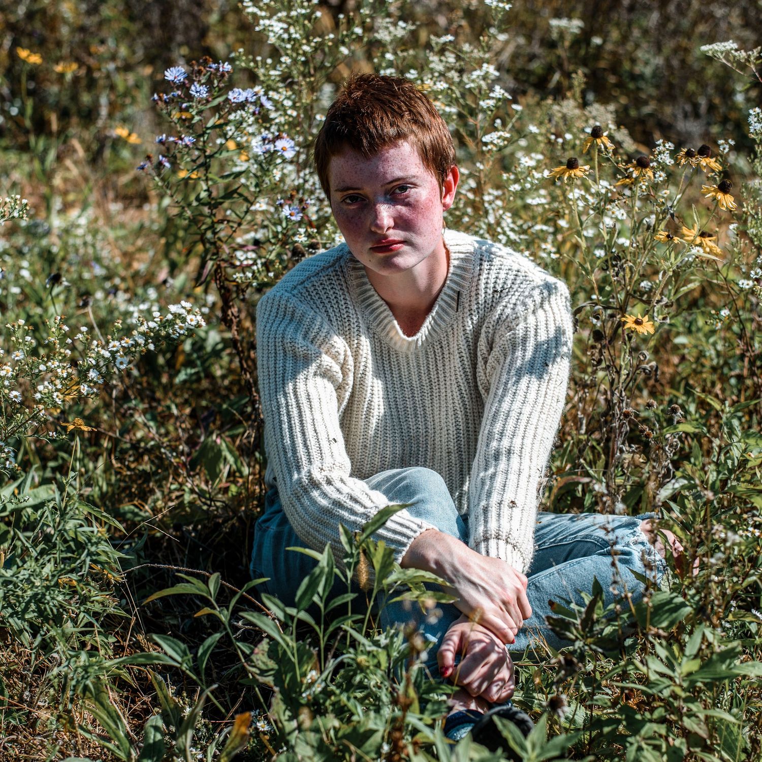 Young person with red hair sitting in a field wearing a cream knit sweater.