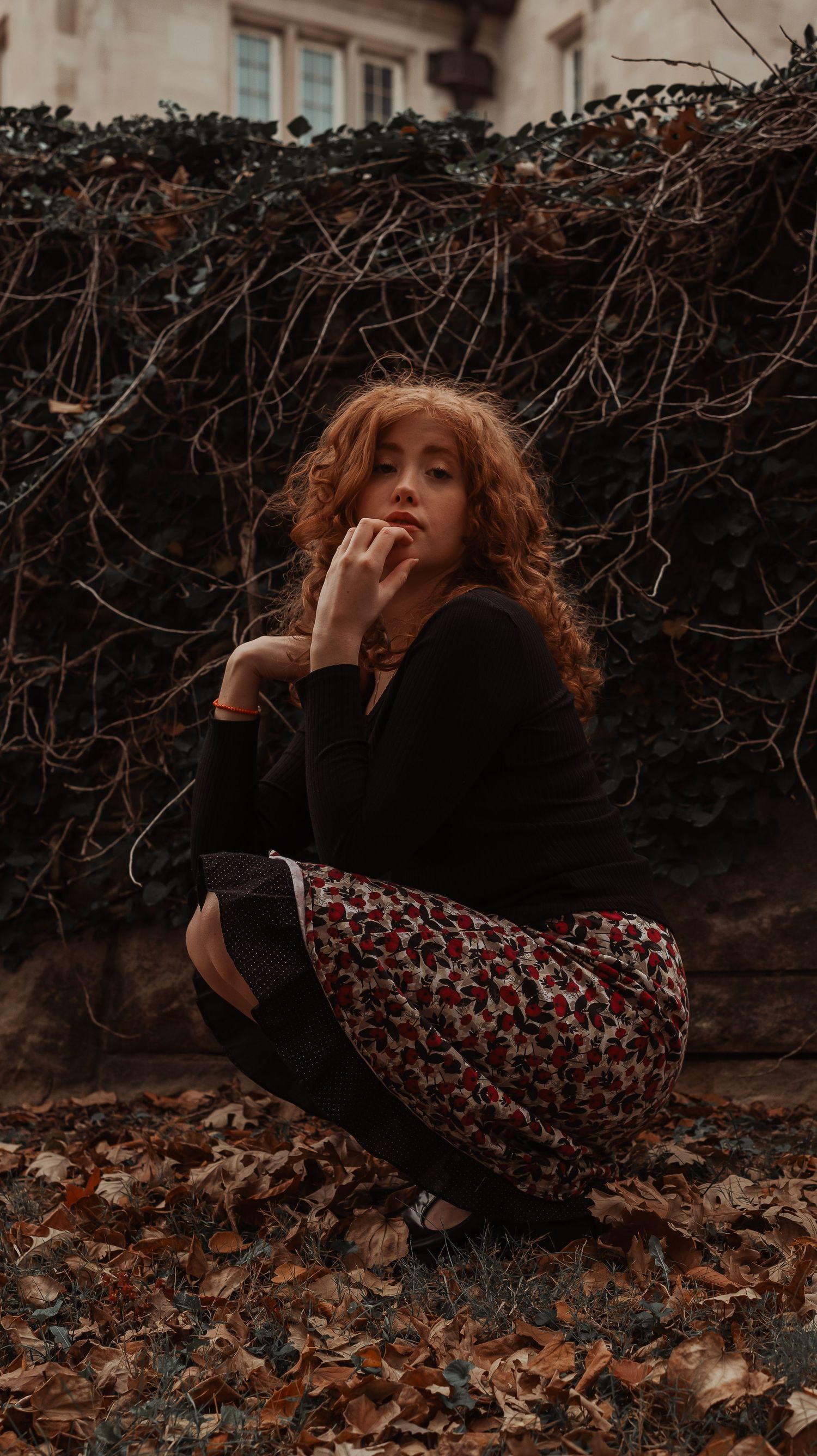 Redheaded woman in black top and floral skirt crouches among fallen leaves by ivy wall.