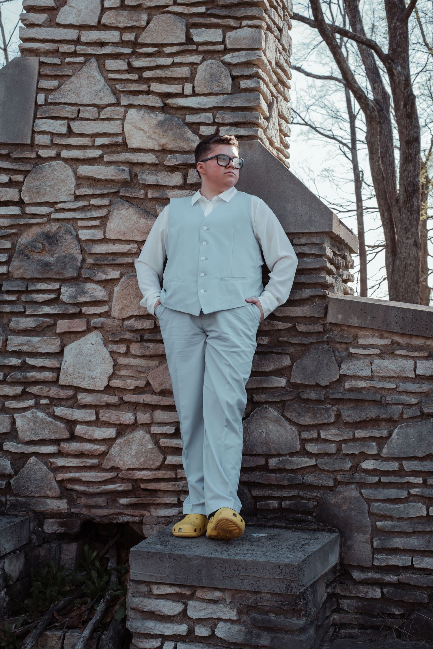 Young man in light blue prom suit with yellow Crocs poses against stone wall outdoors.