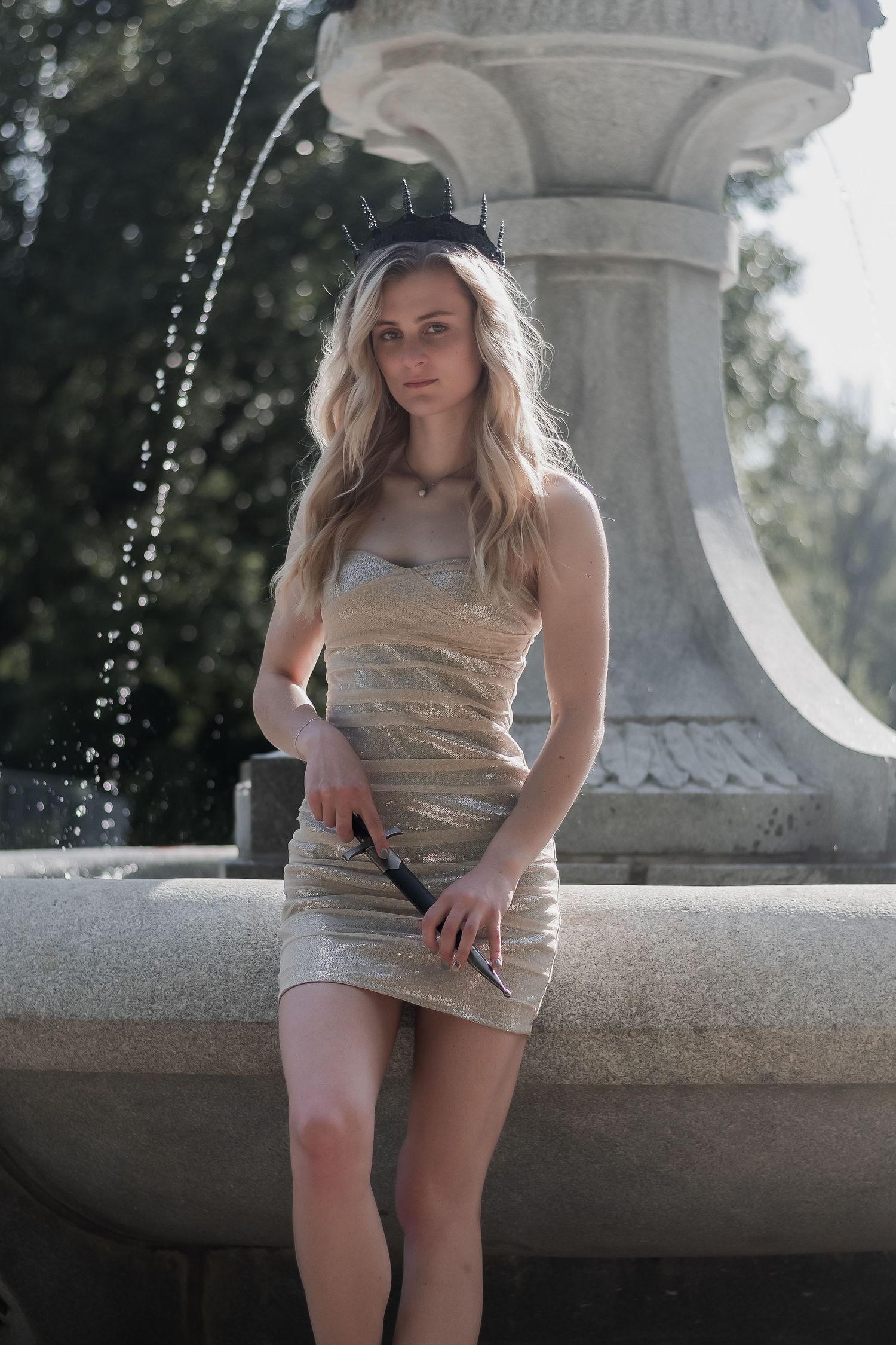Blonde woman in gold bandage dress holds sword beside ornate stone fountain outdoors.