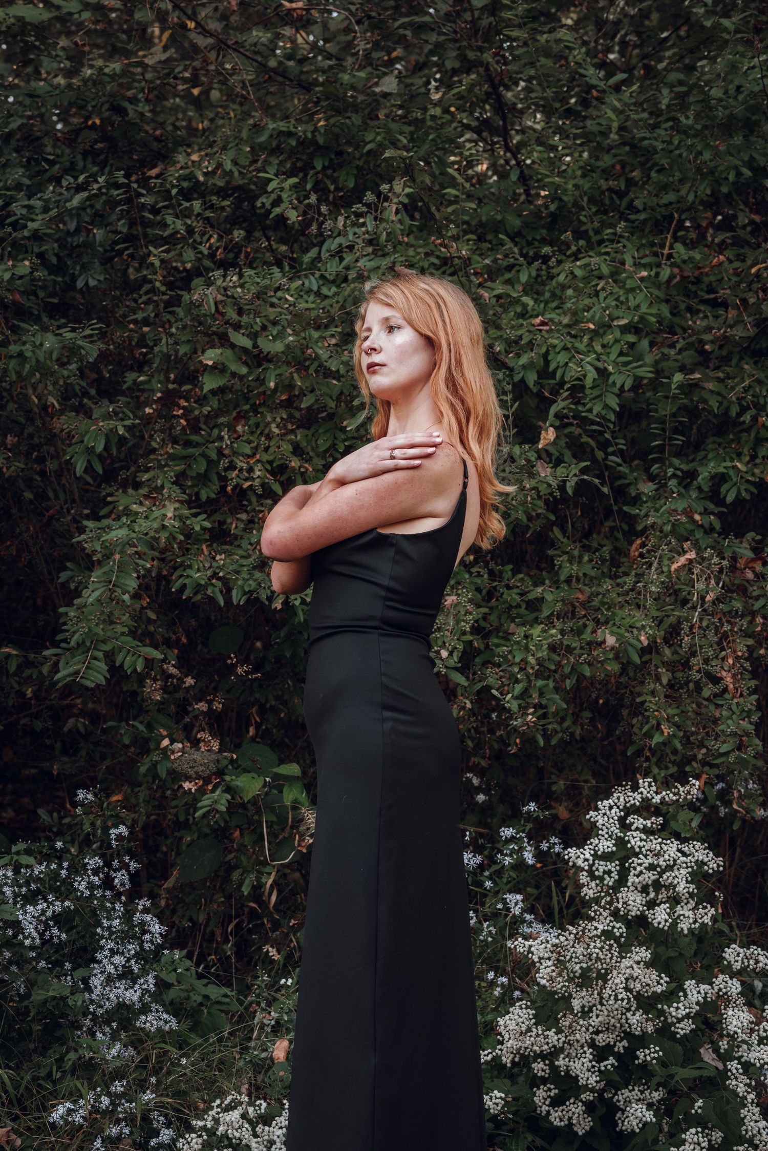 Redhead woman in sleek black maxi dress posing gracefully among green trees and white wildflowers.