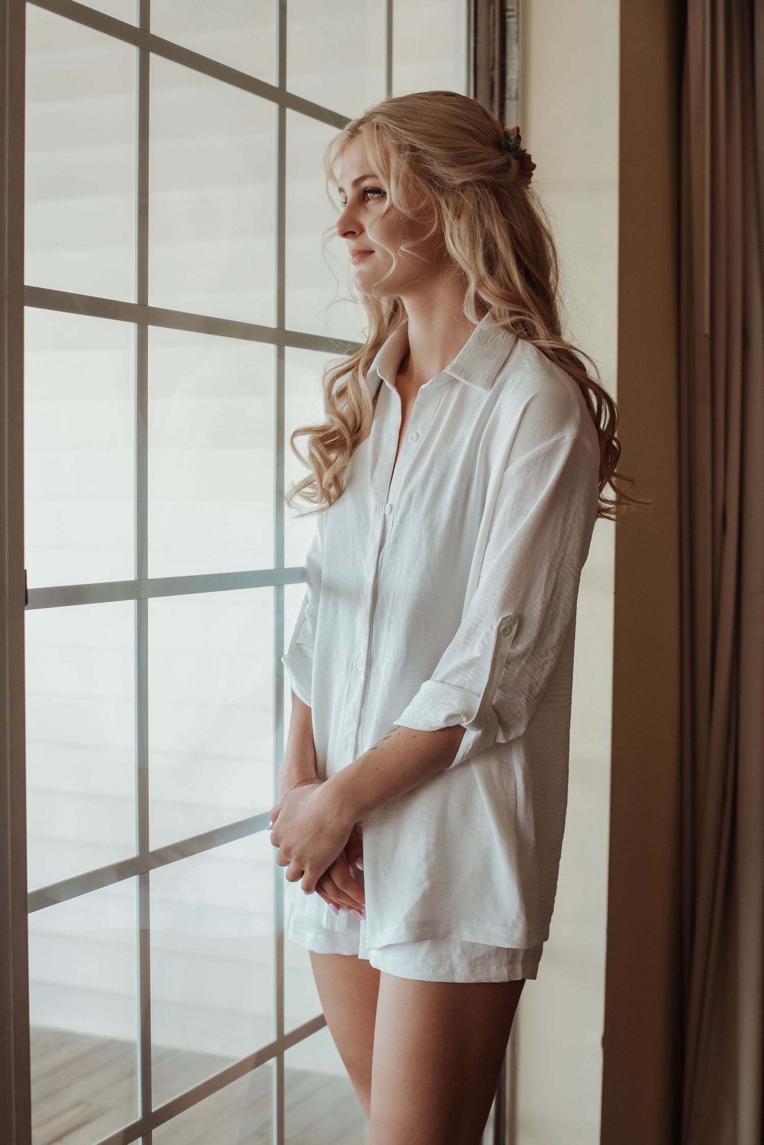 Elegant blonde bride in white oversized shirt gazing out large window during wedding morning prep.