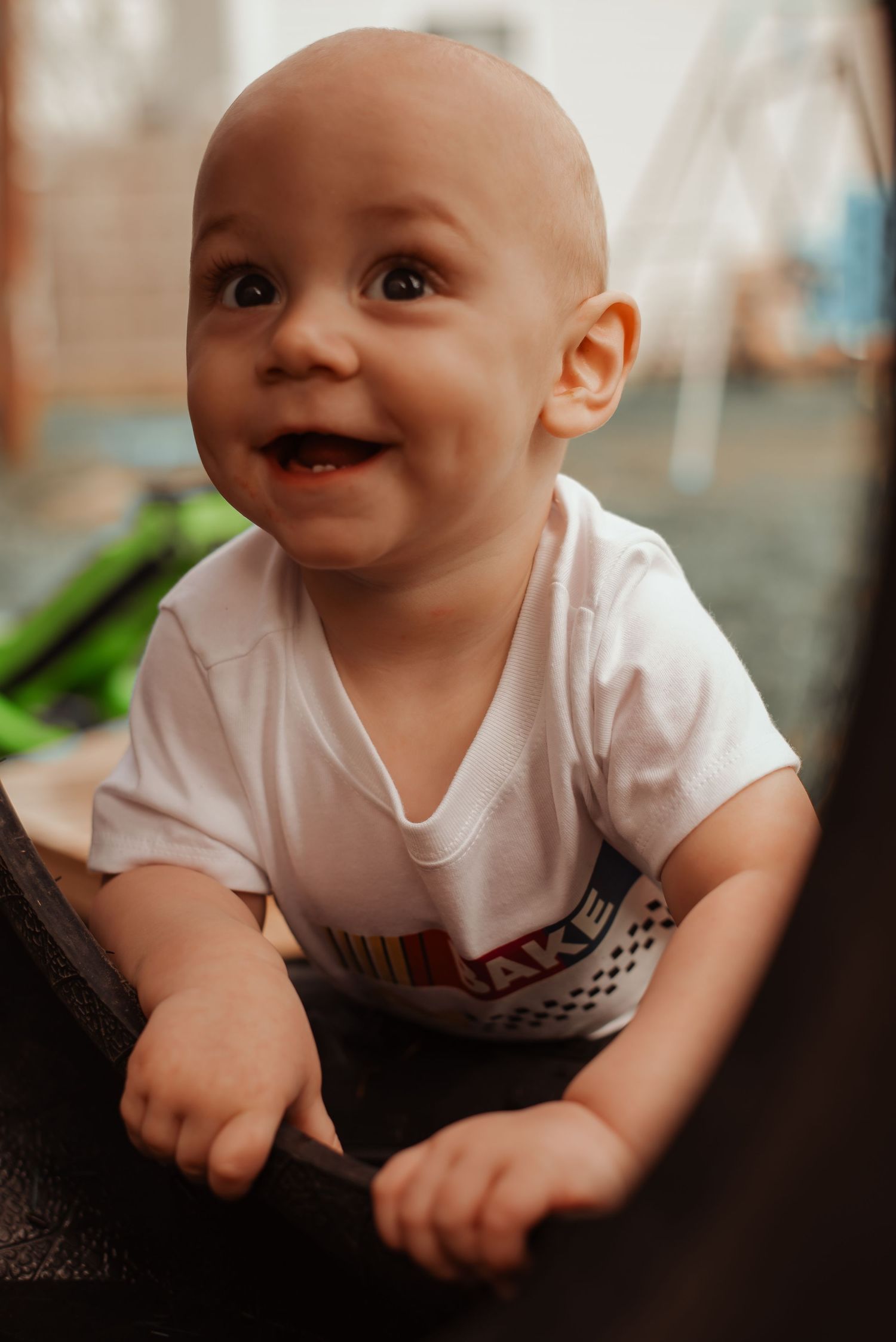 Happy laughing baby in white racing shirt leans on a black tire outdoors.