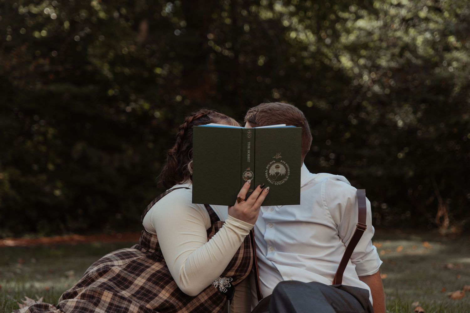 A couple sits outdoors in a park kissing behind a green hardcover book together.