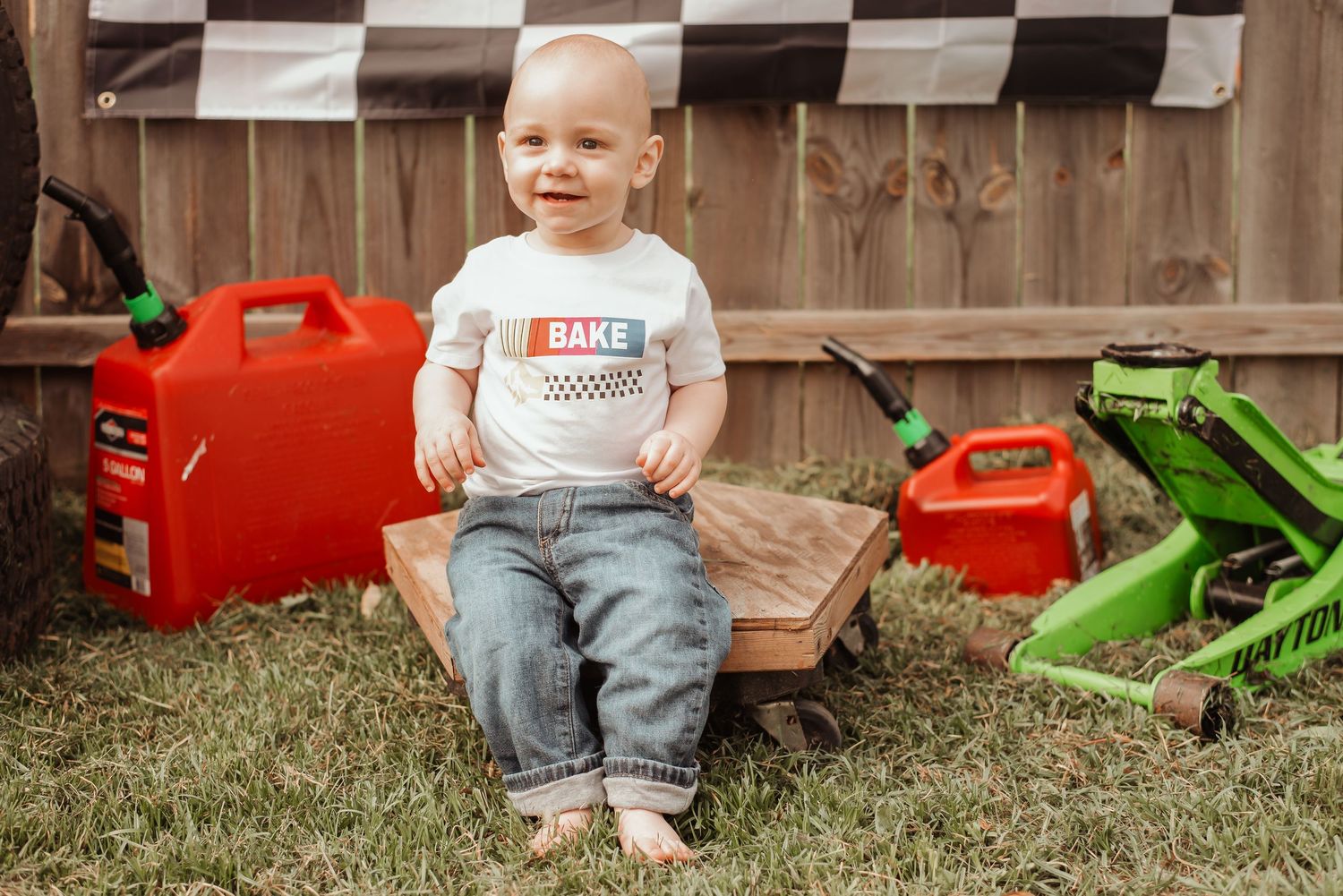 Baby boy in racing onesie sits in wooden box surrounded by red gas cans and toy vehicles outdoors.