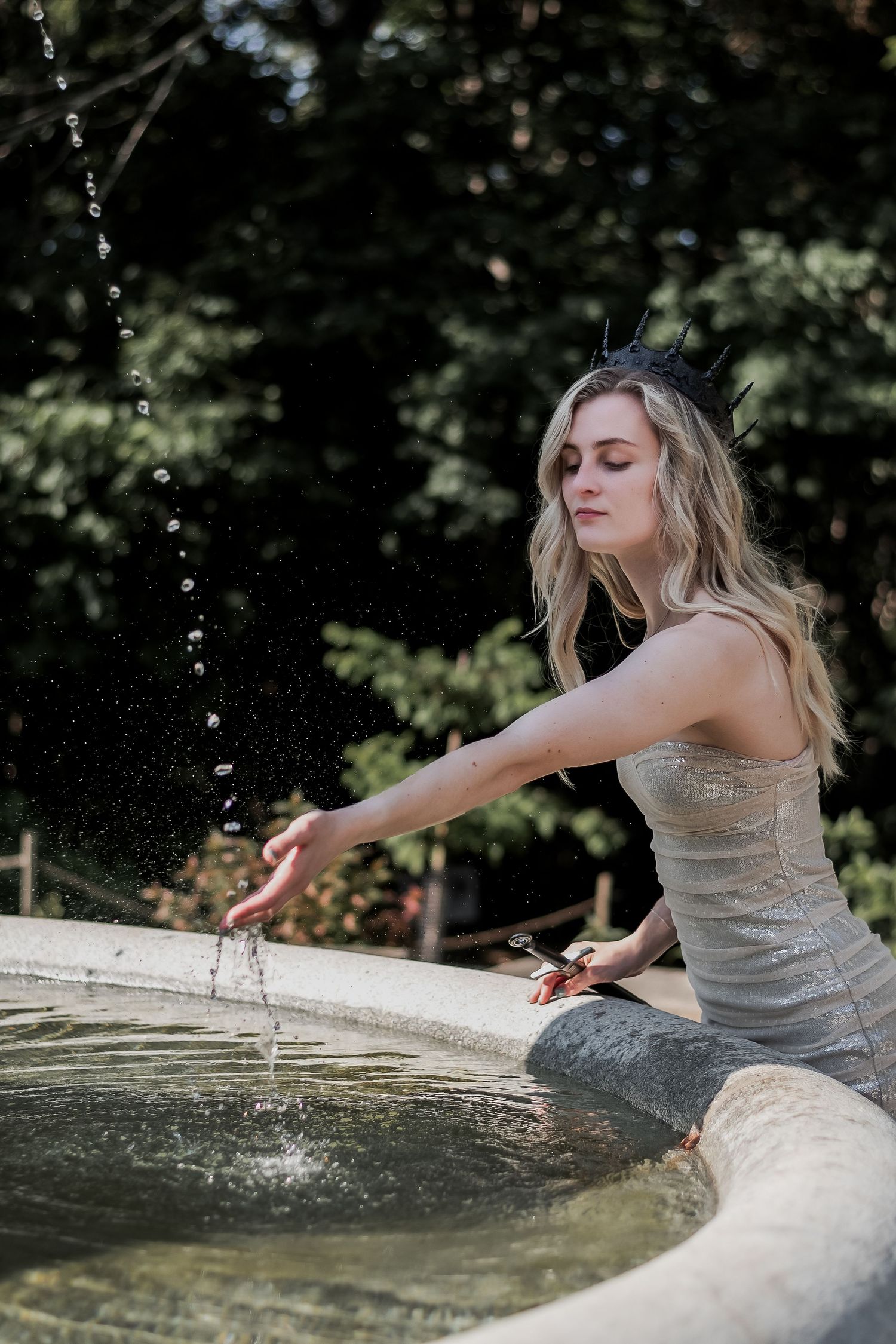 Blonde woman in silver dress sitting on stone fountain splashing water in sunny garden.