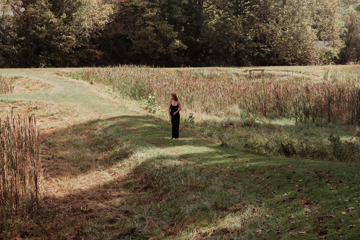 Woman in black outfit stands alone in a vast open meadow surrounded by tall grass and trees.