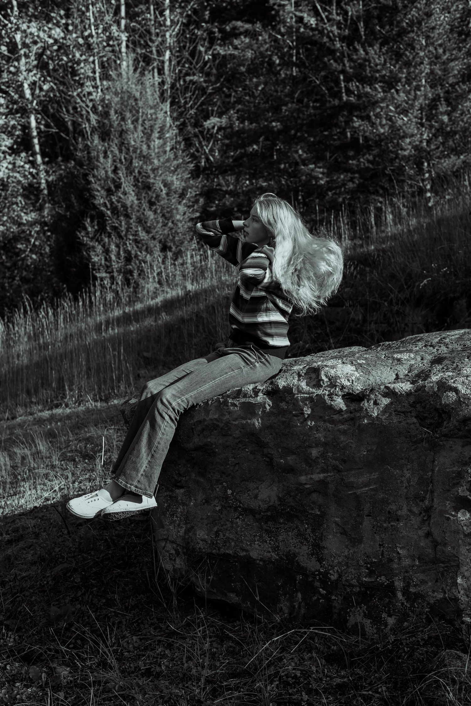 Black and white photo of blonde woman sitting on rock photographing nature in forest.